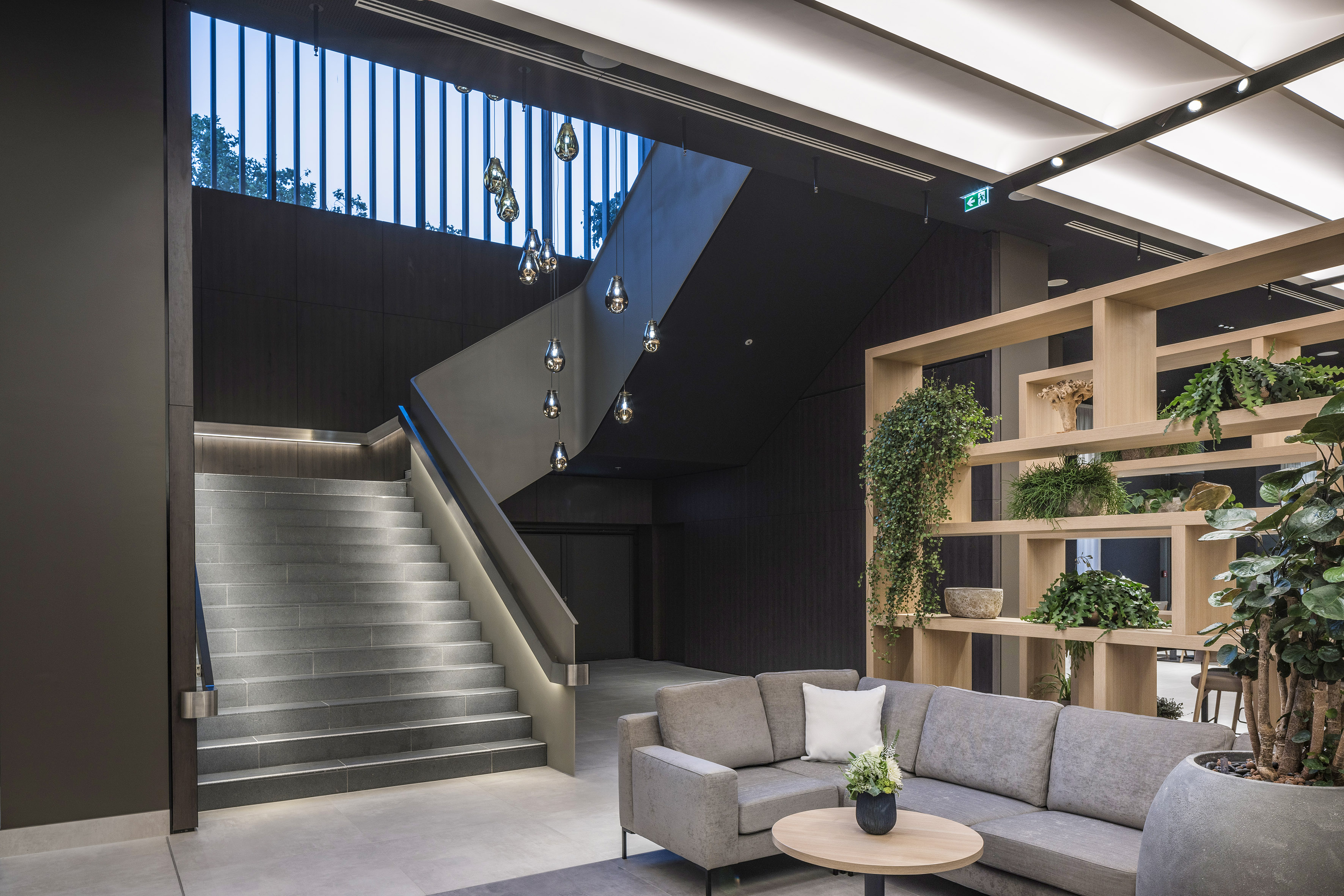 Modern office lobby, illuminated staircase, gray sofa, and green plant display.