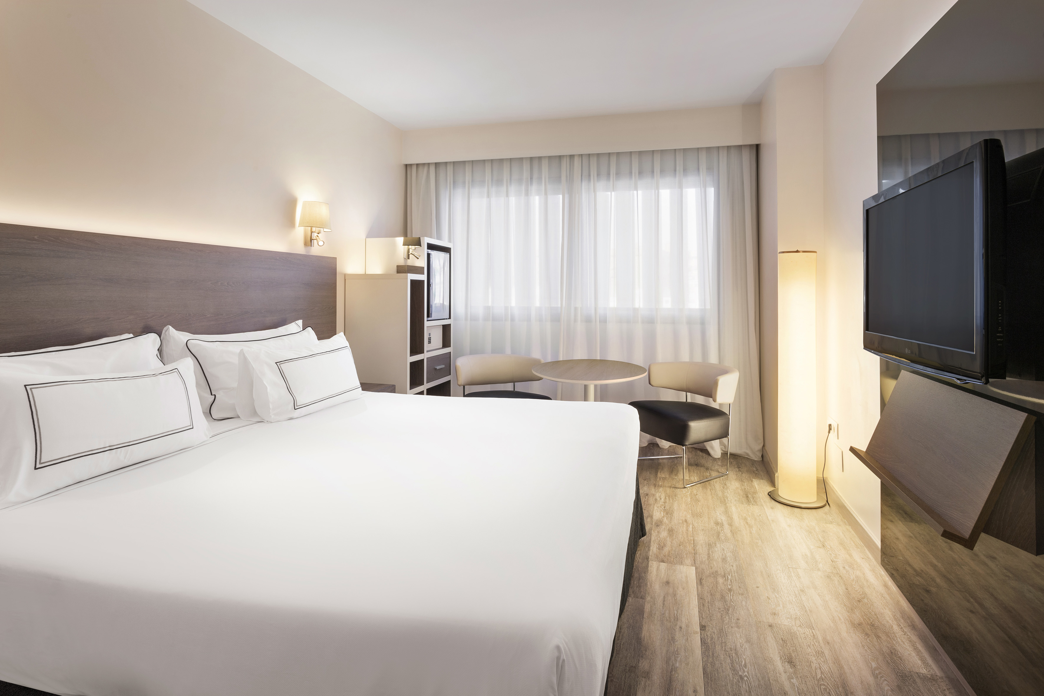 Modern hotel room with large bed, sheer curtains, wood floor, TV, and seating area.
