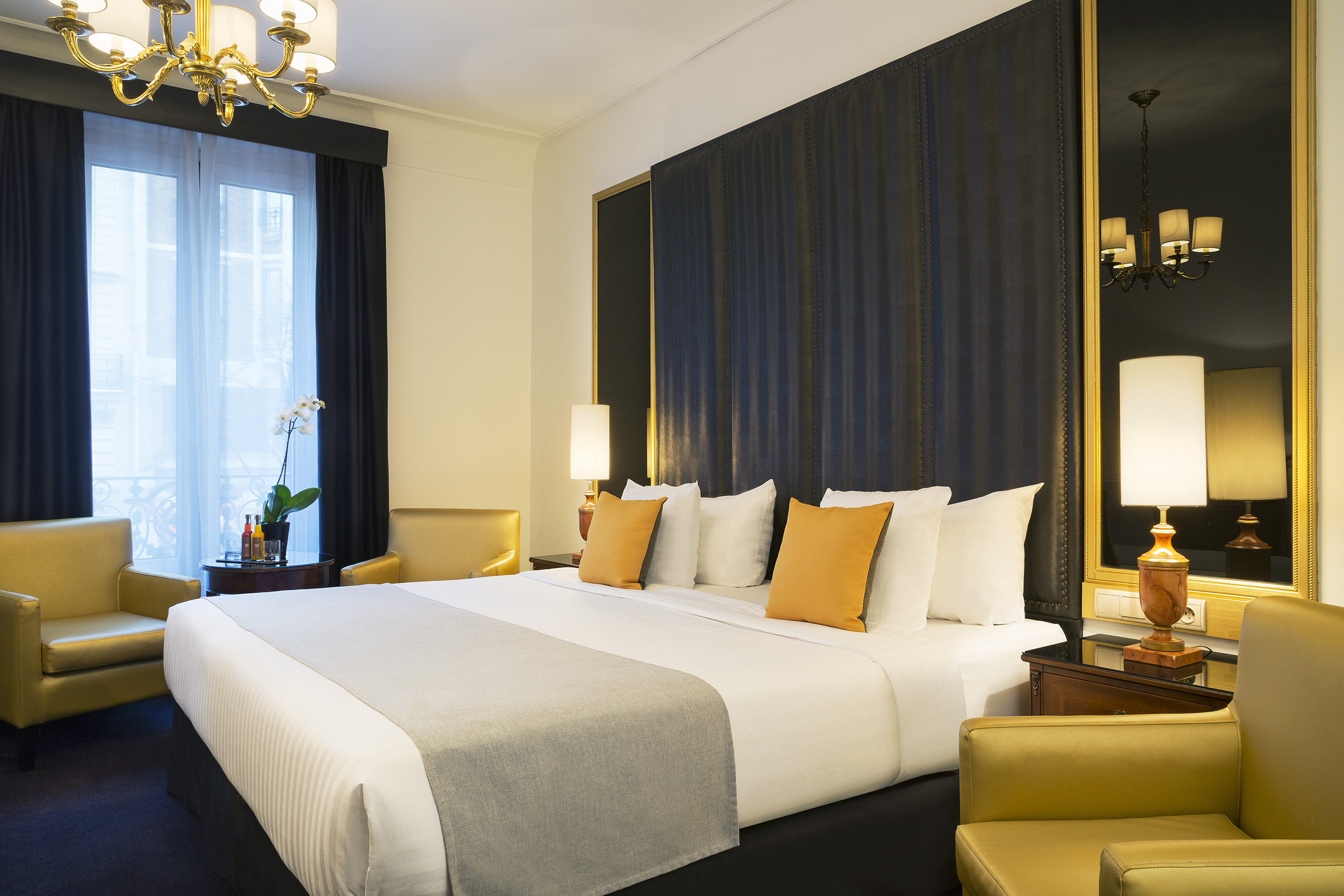 Elegant hotel bedroom with plush bed, gold armchairs, and dark headboard.