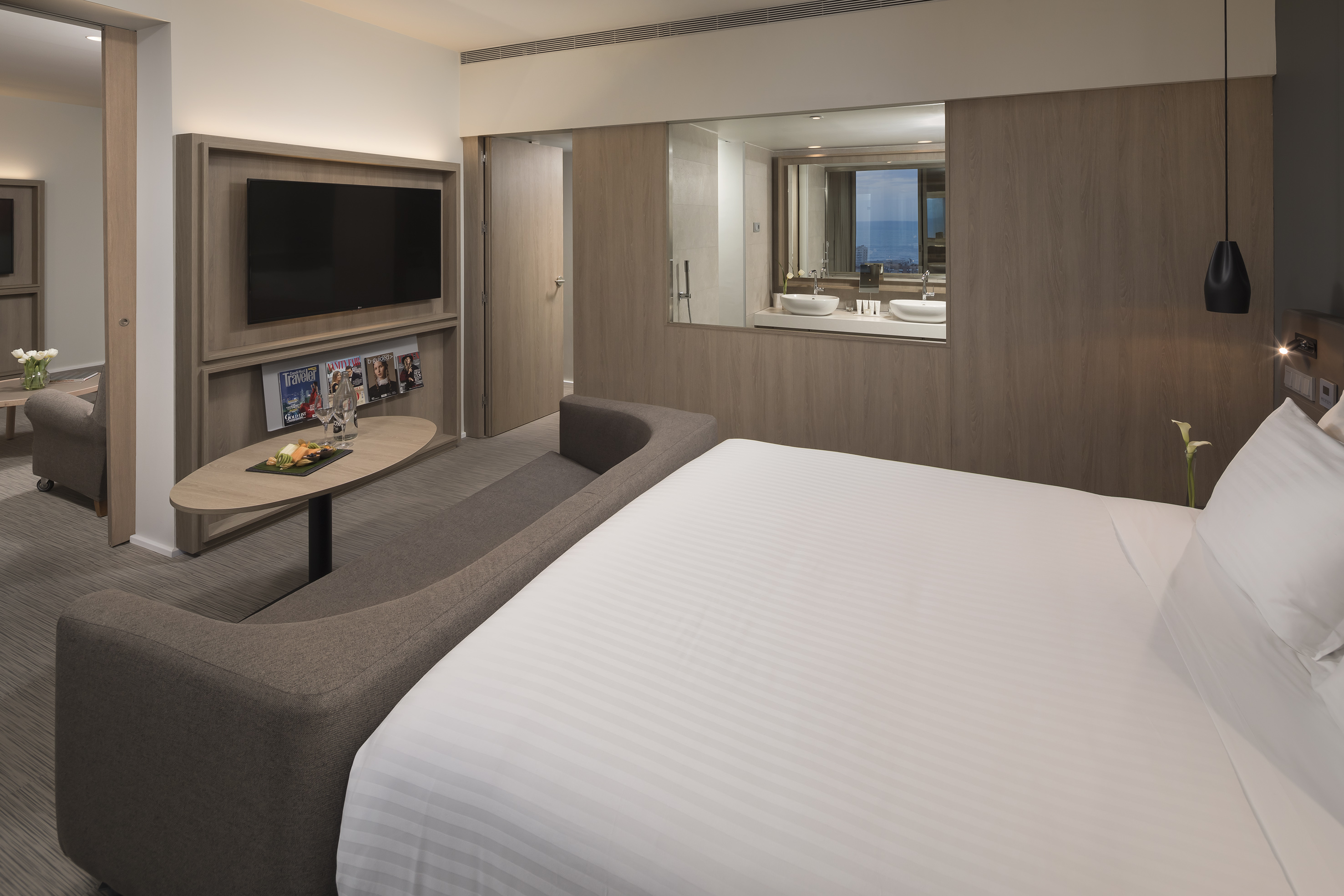 Modern hotel suite with bed, TV, lounge area, and bathroom reflecting sea view.