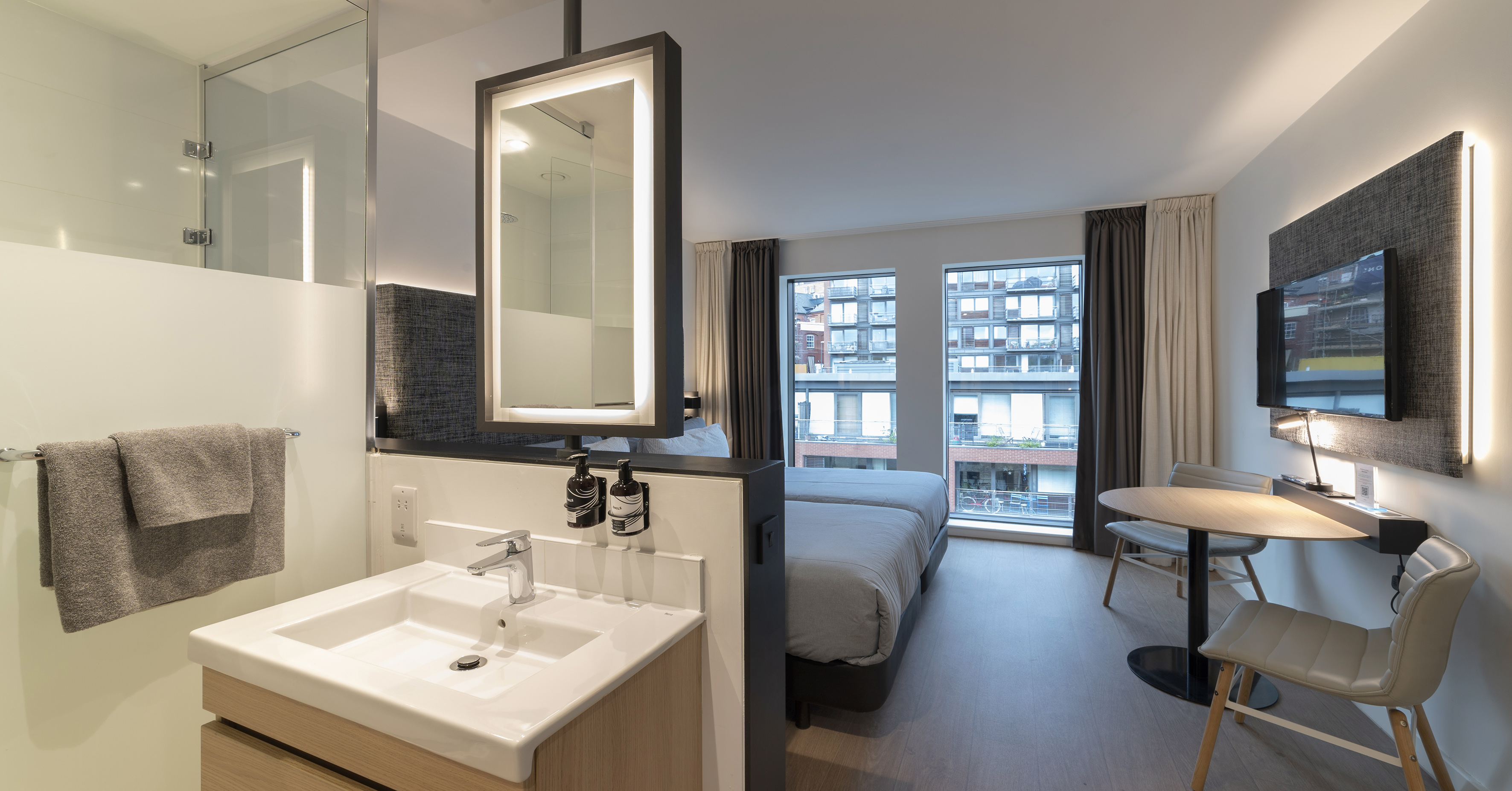 Contemporary hotel room with twin beds, open bathroom, and urban city view.