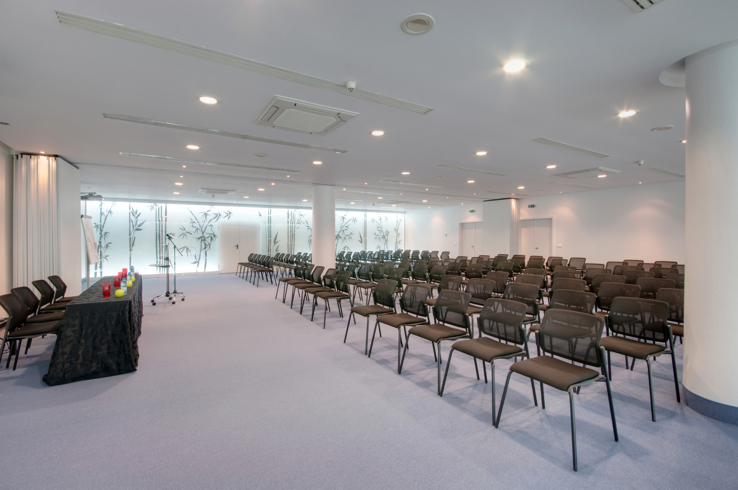 Empty conference room with rows of black chairs, presentation table, and bamboo decor.