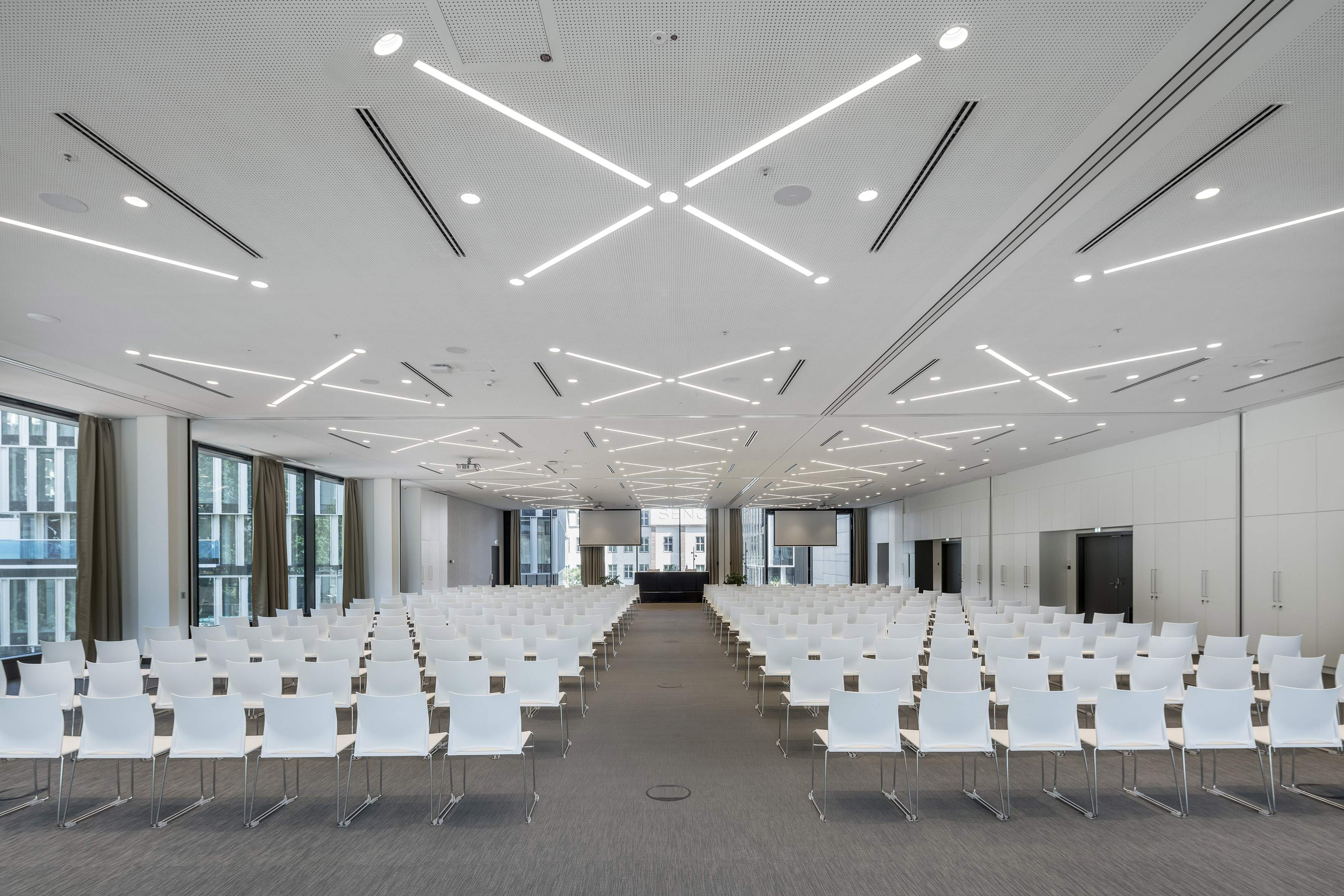 Modern conference hall with white chairs, dual screens, and geometric ceiling lights.
