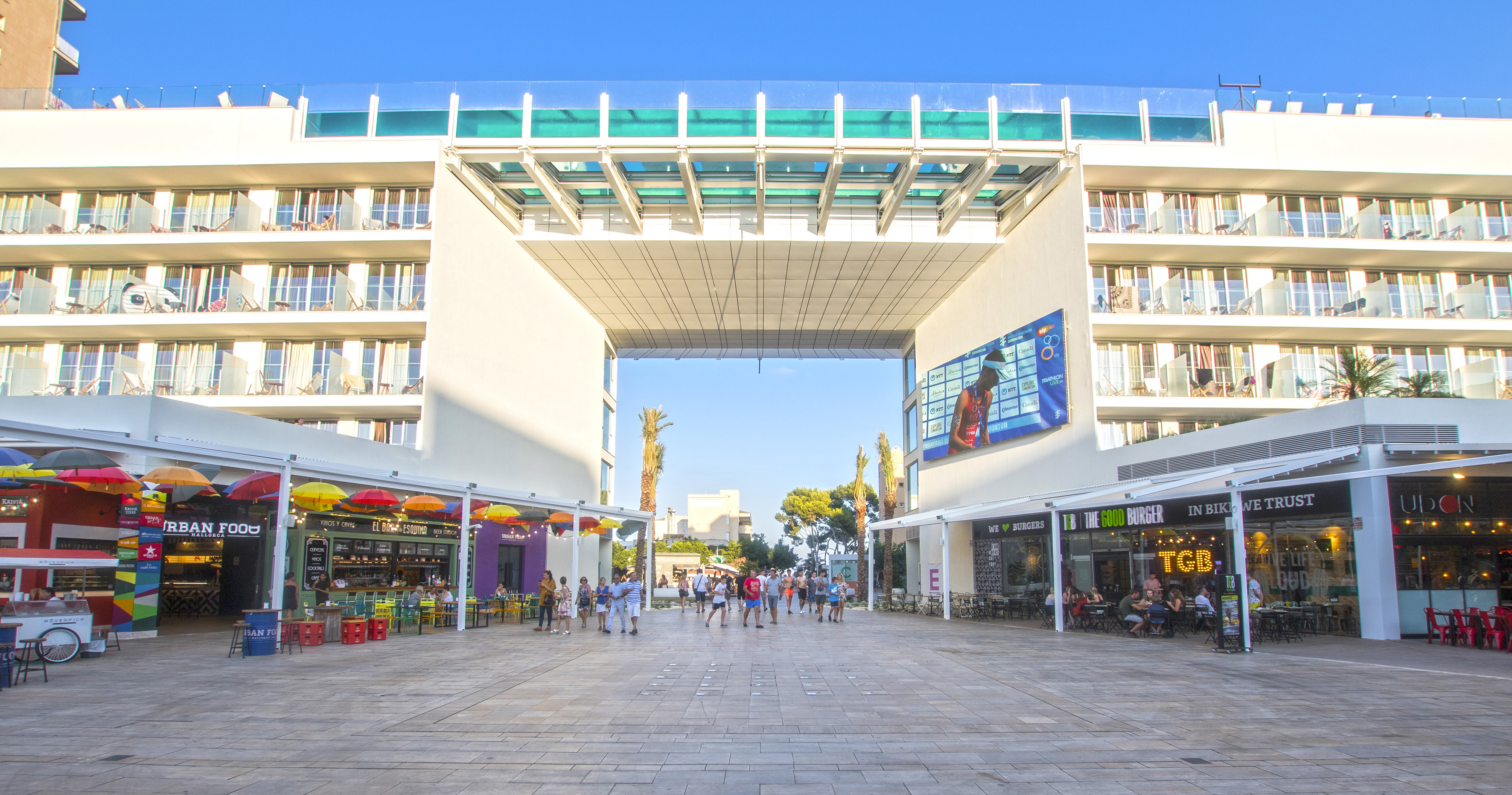 Modern resort hotel with archway, rooftop pool, and lively outdoor dining plaza.
