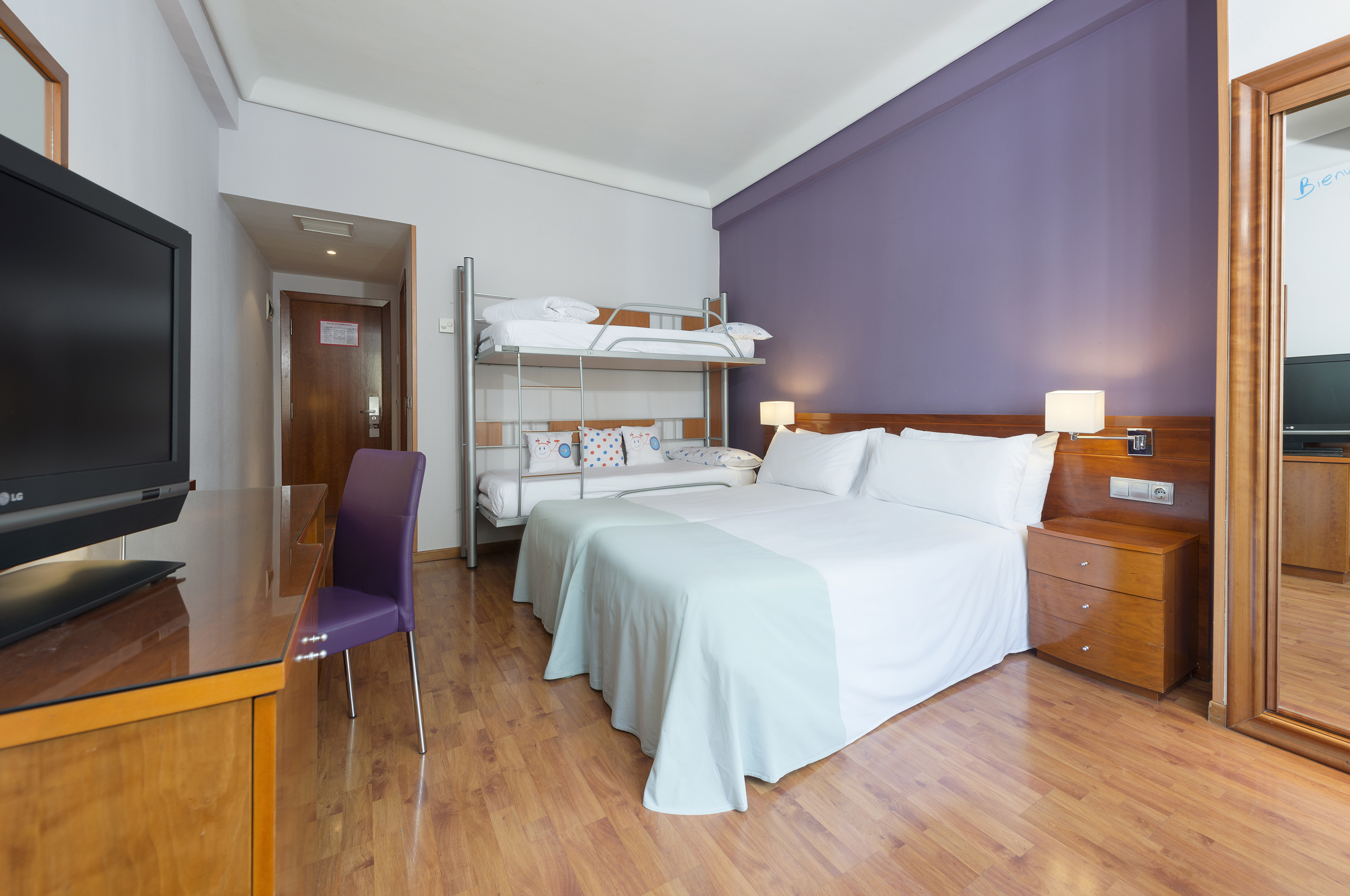 Modern hotel family room with double bed, bunk beds, purple wall, and wooden floor.