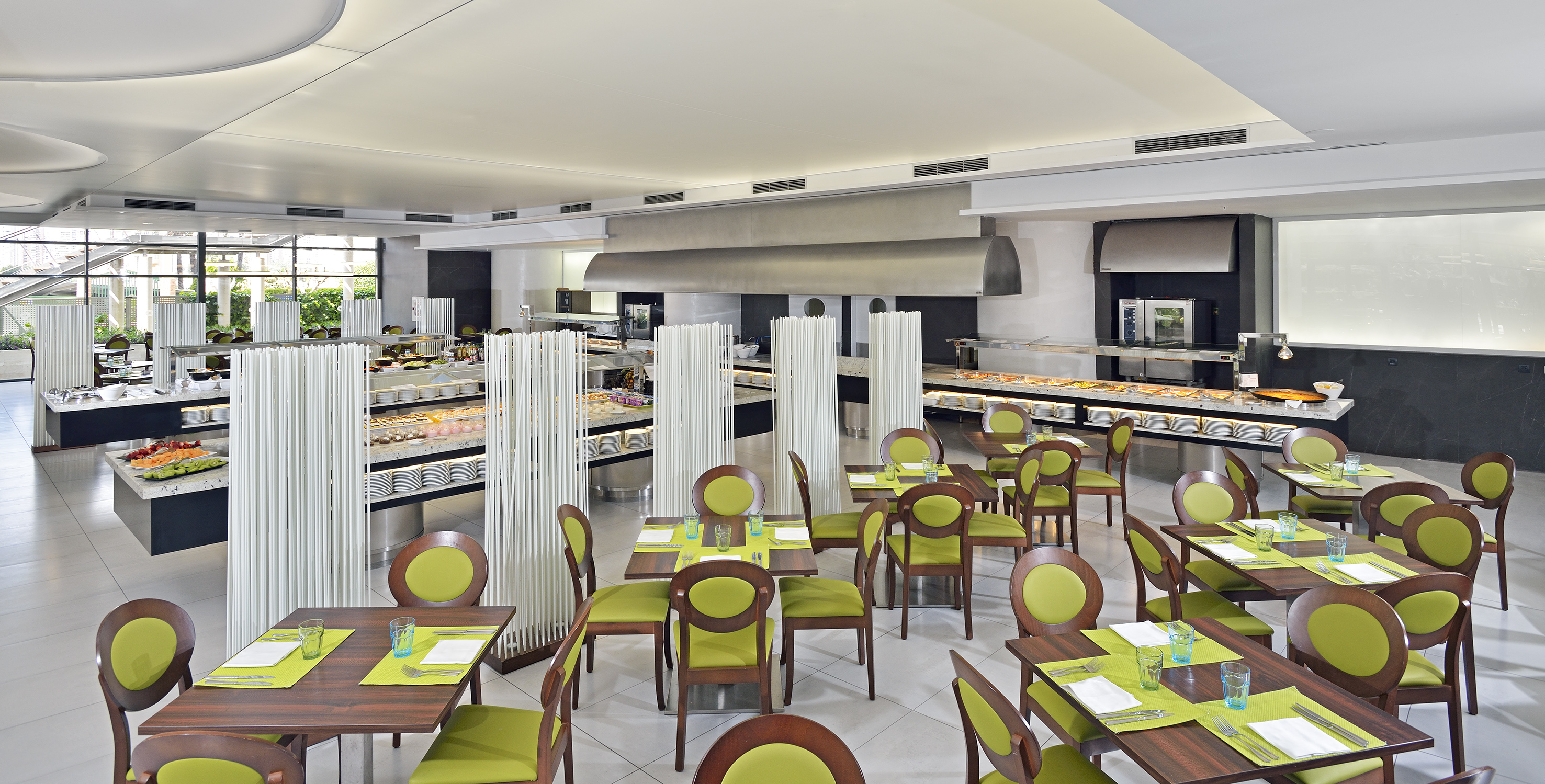 Modern hotel buffet restaurant with green chairs and self-service food stations.