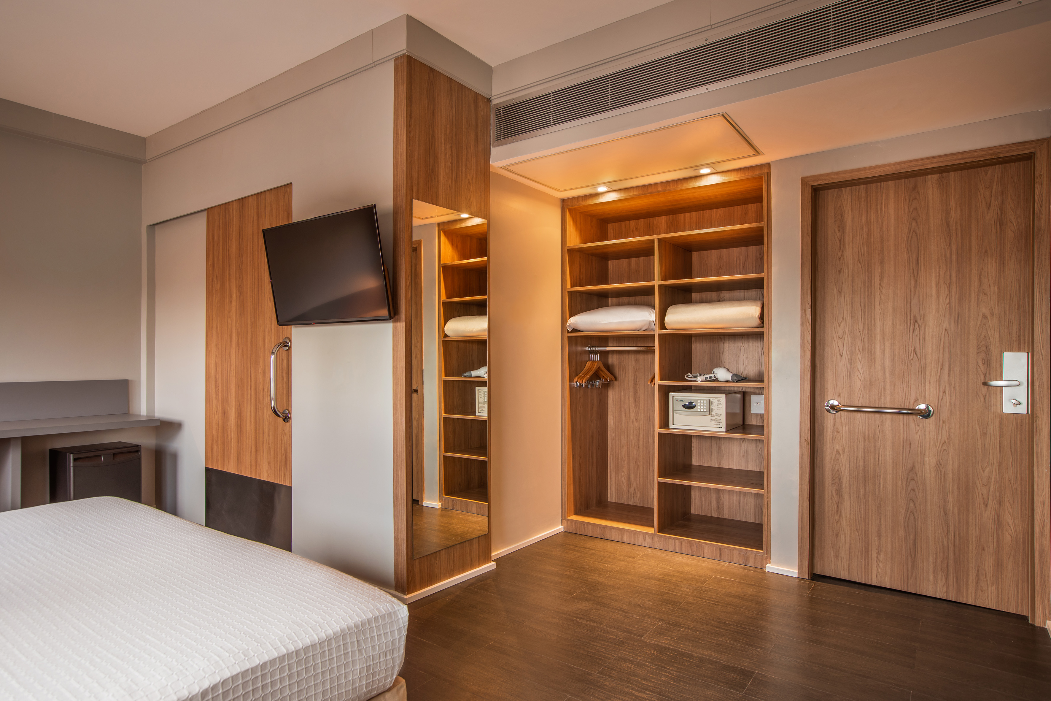 Modern hotel room with open wooden wardrobe, wall TV, and bed.