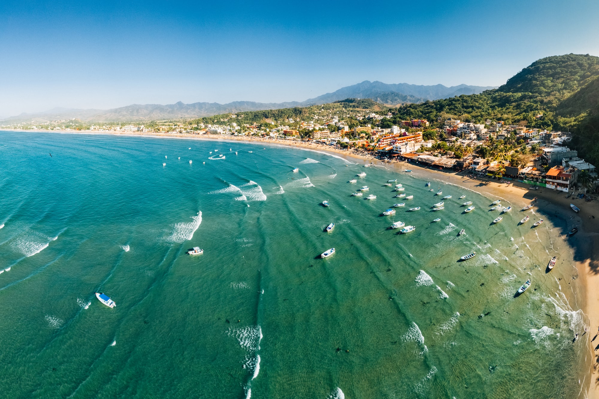Panorámica aérea de playa, bahía con barcos, pueblo costero y montañas.