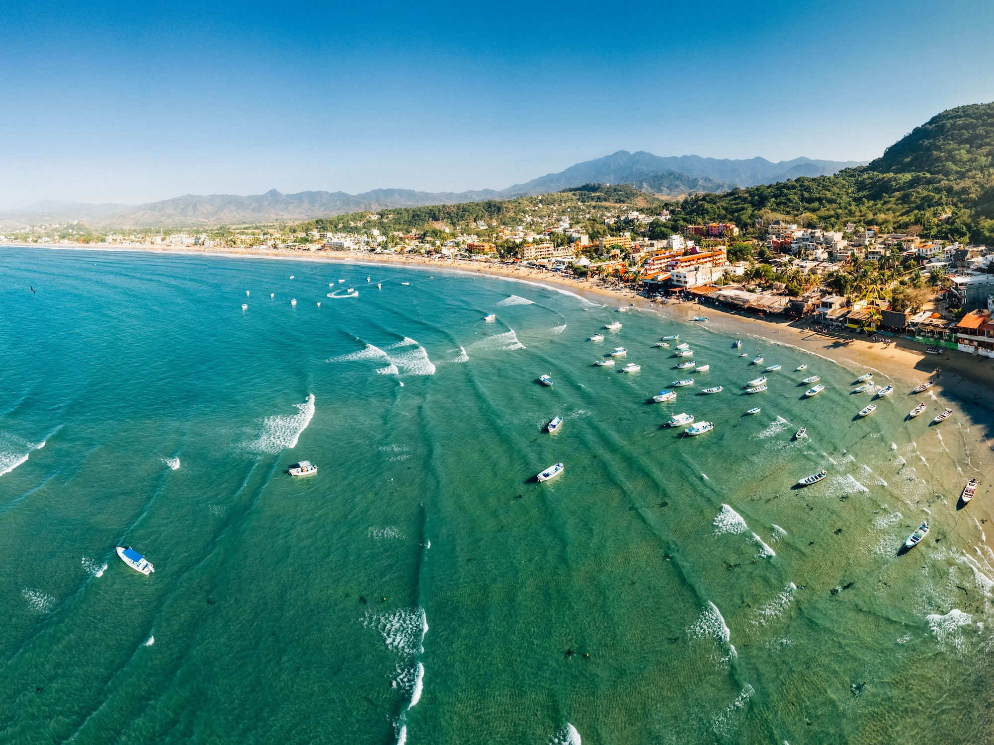 Aerial view of a coastal town bay with boats, sandy beach, and green mountains.