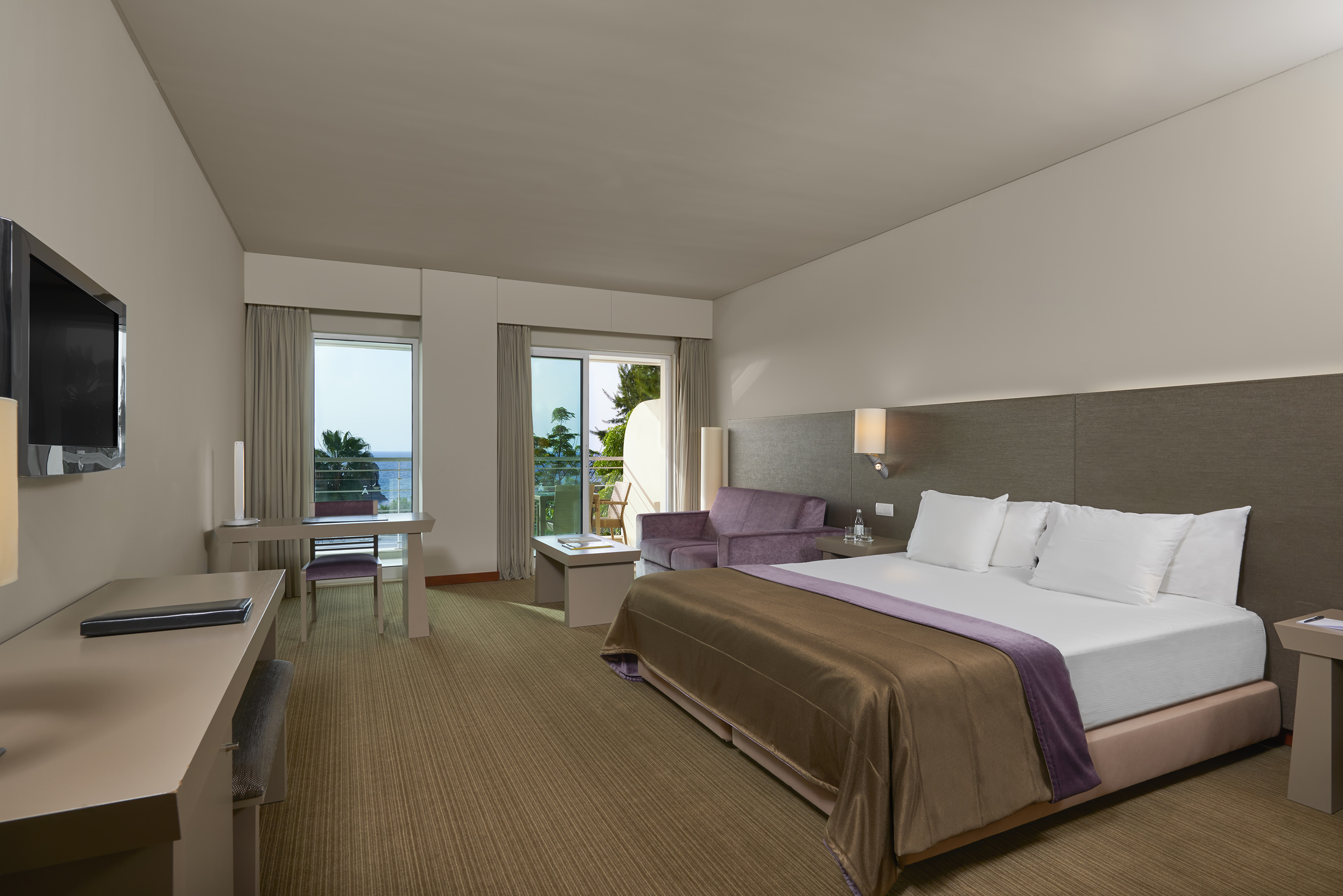Modern hotel room with king bed, balcony, and distant sea view.