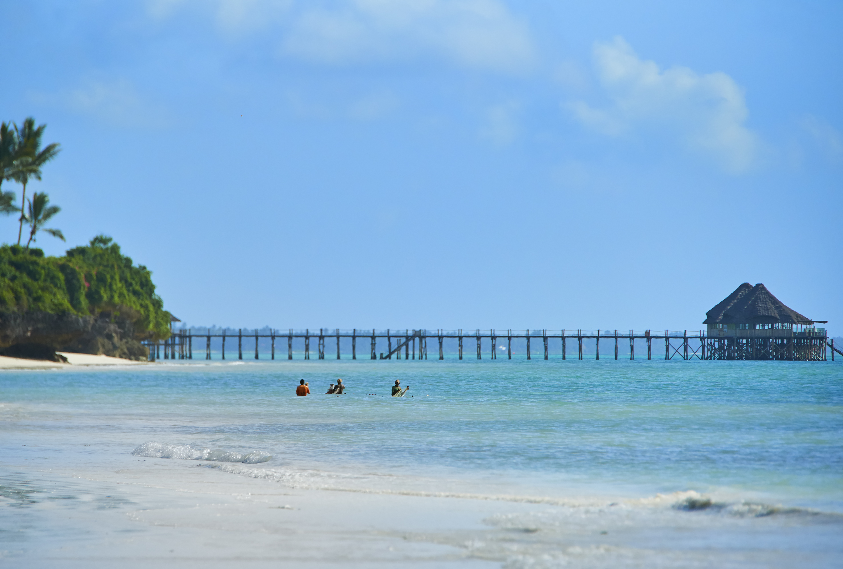 Tropical beach: swimmers, long pier to overwater restaurant, lush island.