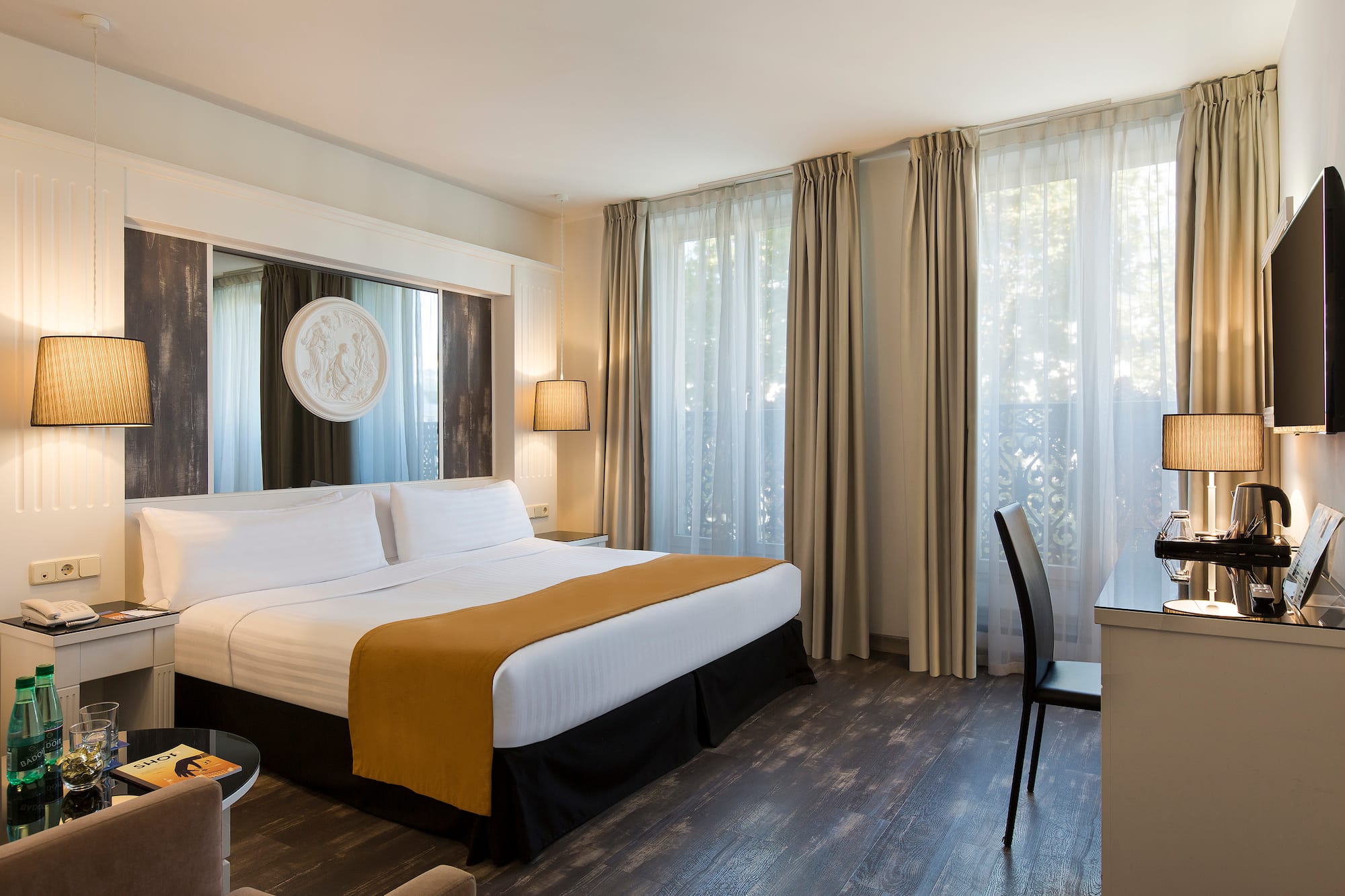 Elegant hotel guest room with king bed, desk, and sunlit windows.