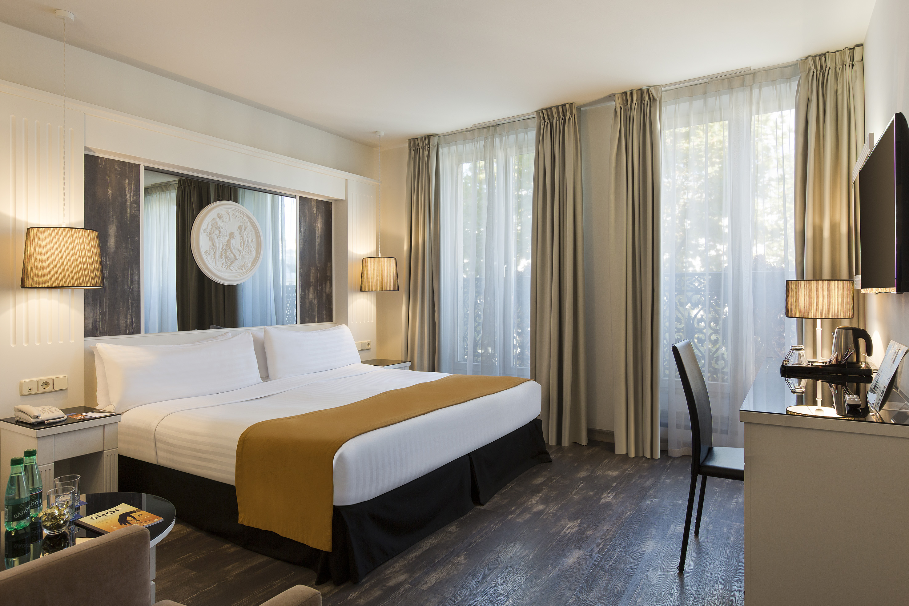 Elegant hotel guest room with king bed, desk, and sunlit windows.