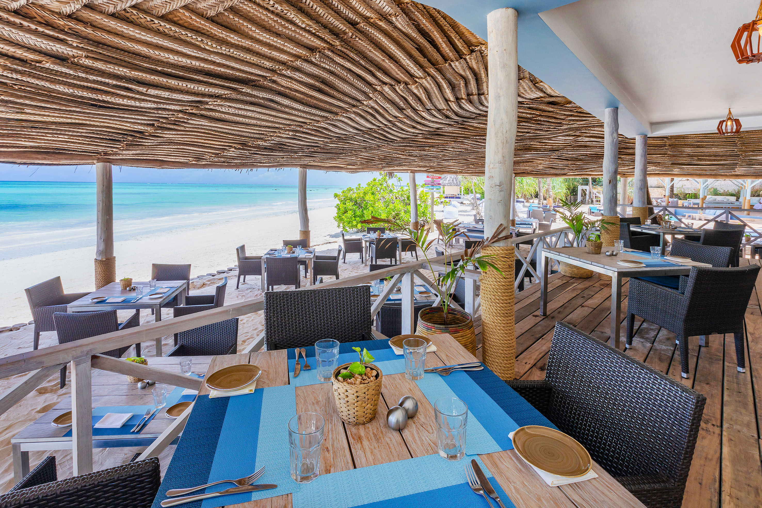 Beachfront restaurant with thatch roof, wooden tables, and turquoise ocean view.