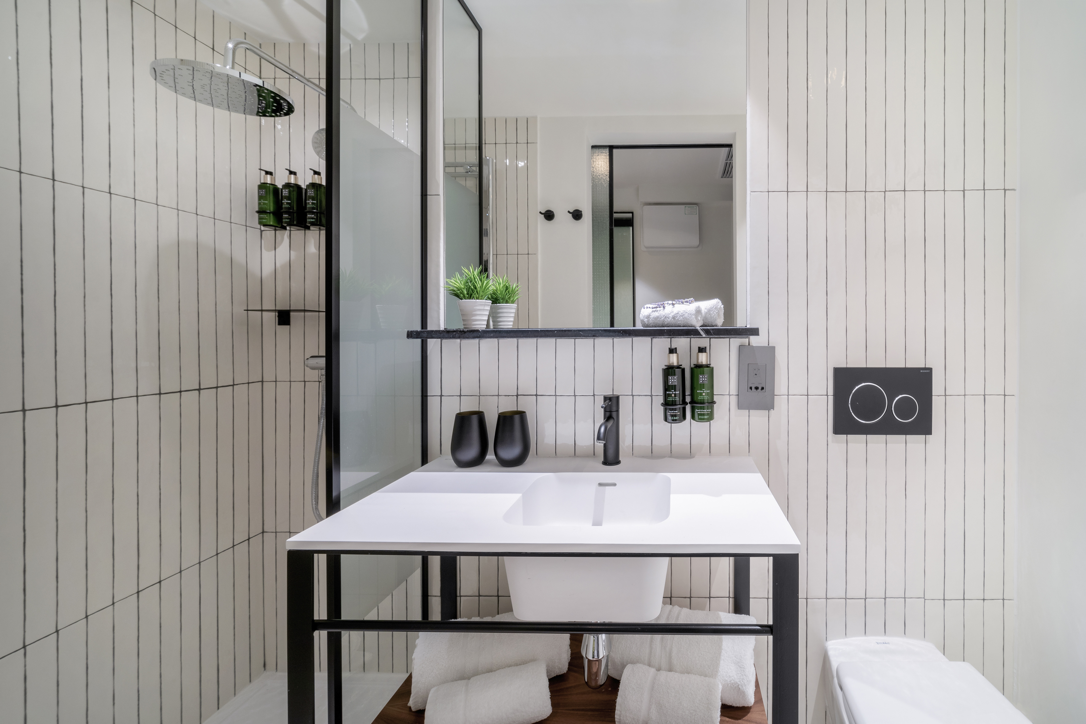 Modern hotel bathroom with white vertical tiles, vanity, rain shower, and black accents.