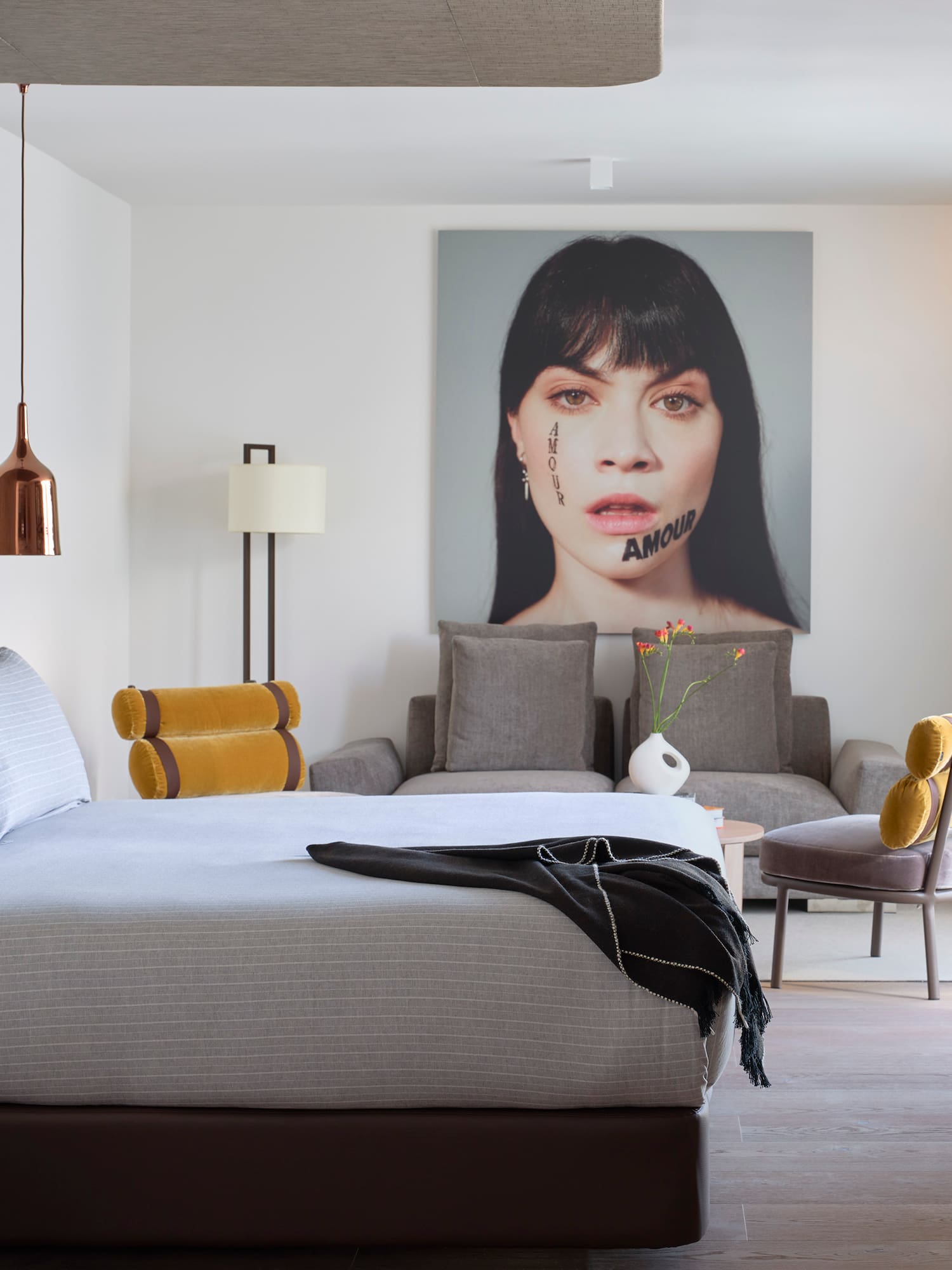 Modern bedroom with striped bed, copper lights, portrait art, gray couch, and plant.