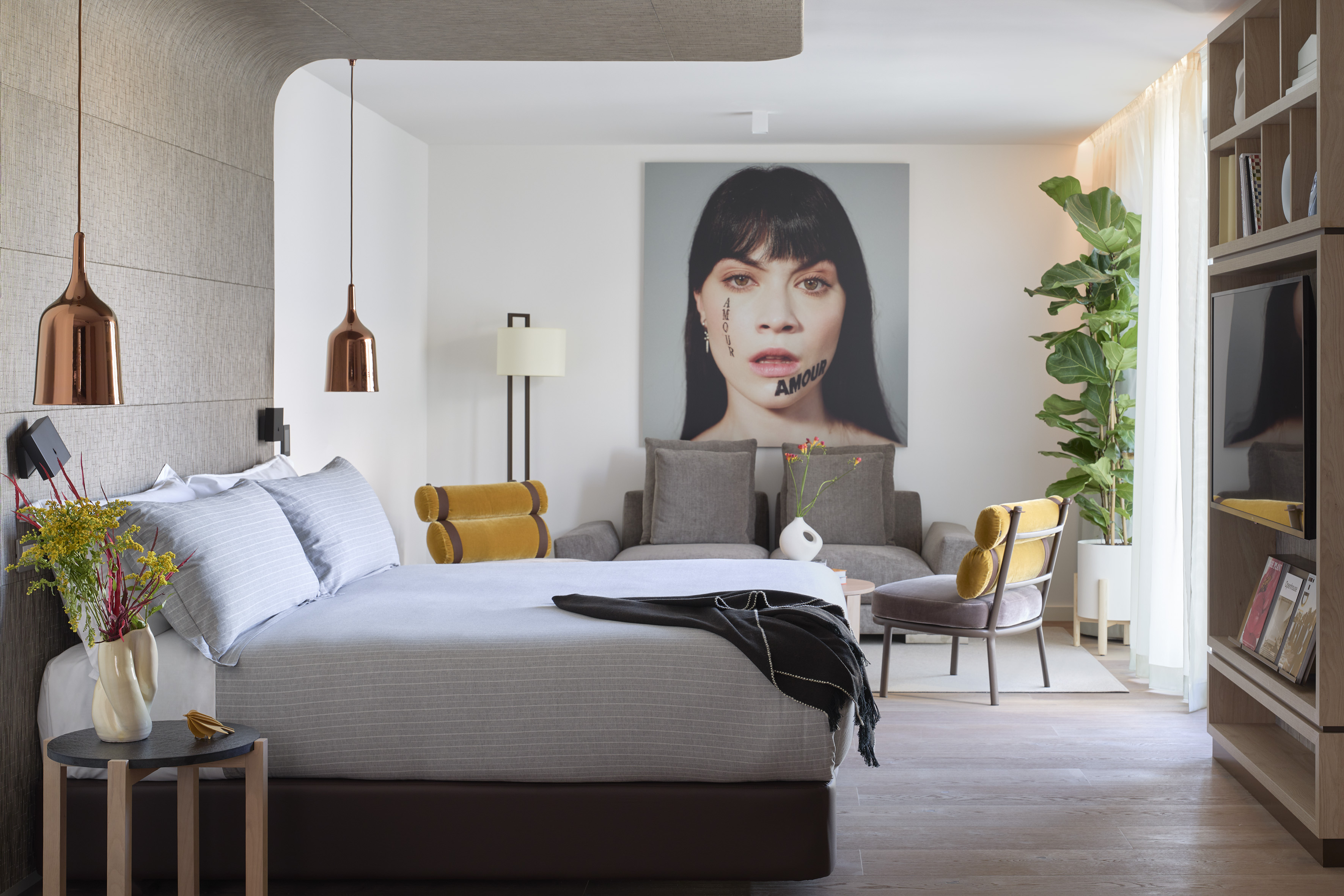 Modern bedroom with striped bed, copper lights, portrait art, gray couch, and plant.