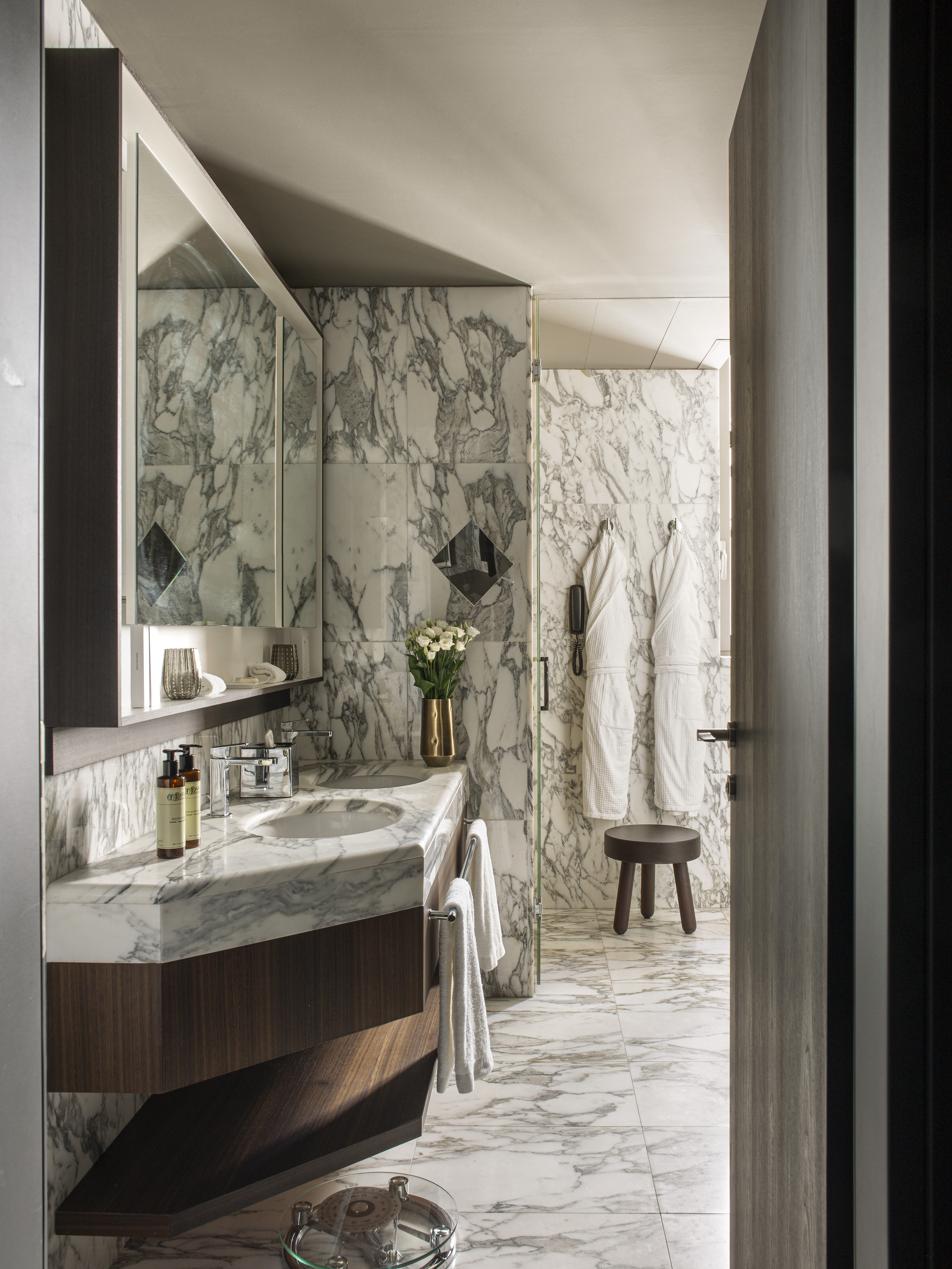 Elegant marble bathroom with double vanity, dark wood, and hanging bathrobes.