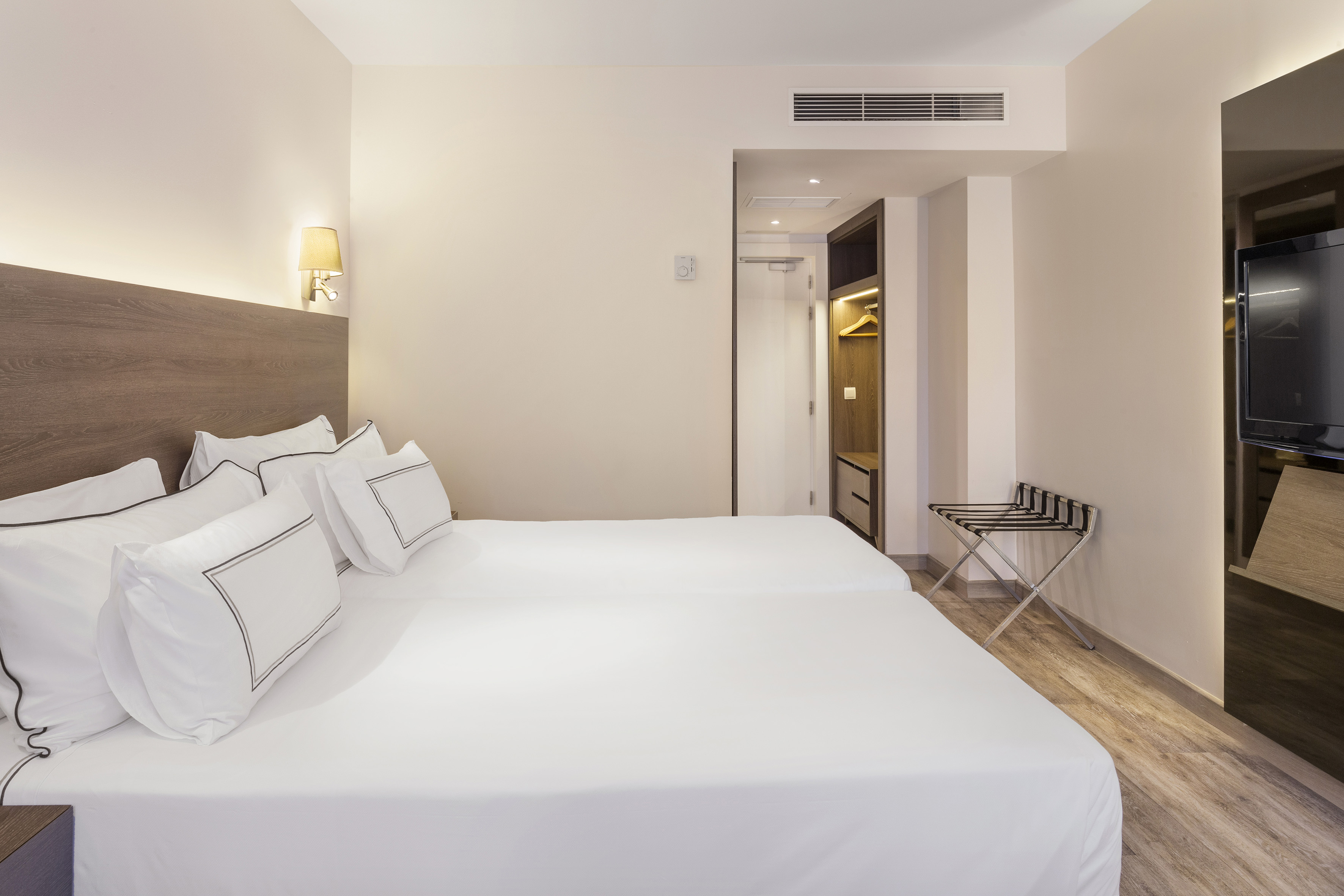 Modern hotel room with two clean twin beds, closet, TV, and luggage rack.