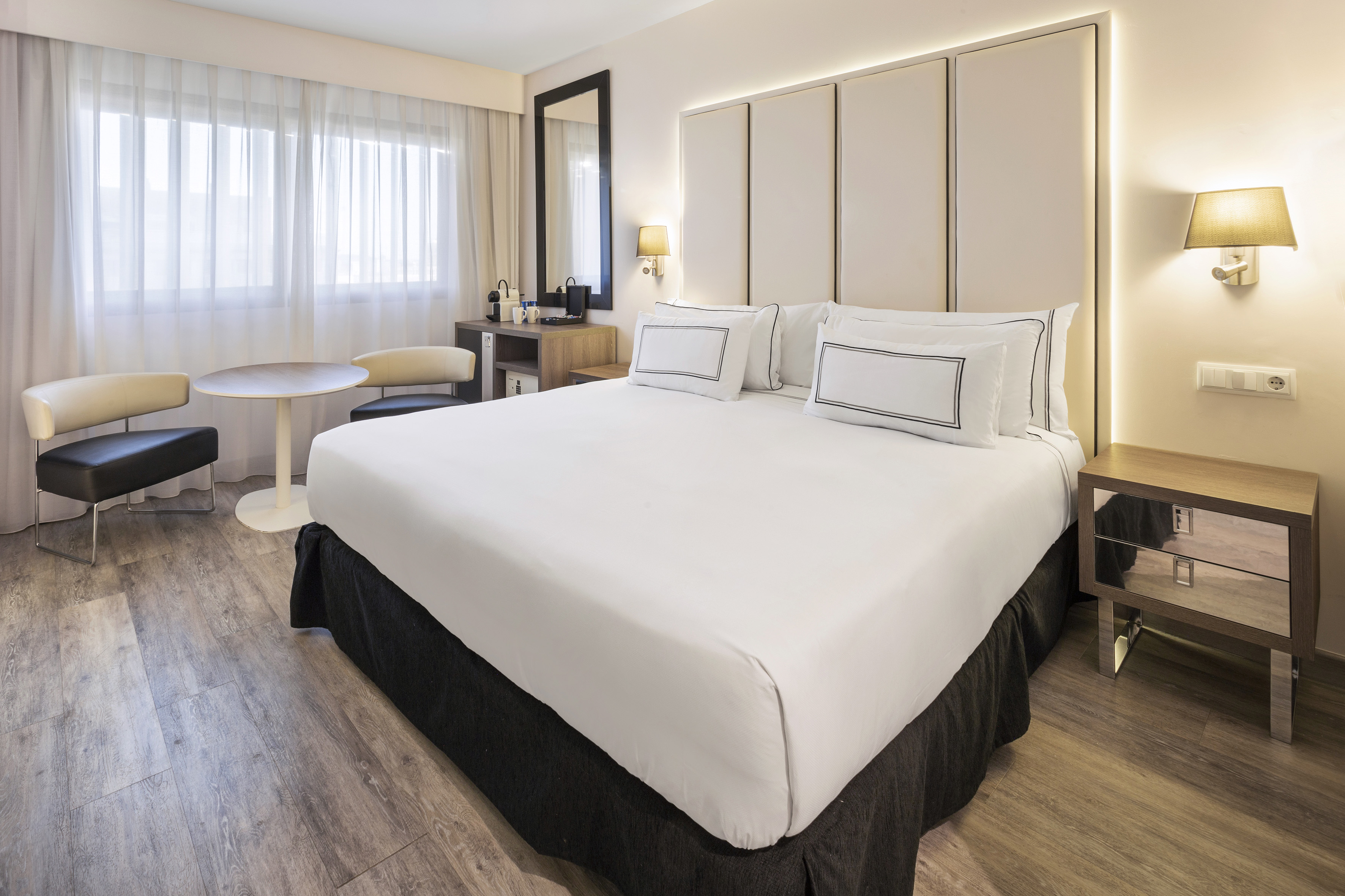 Modern hotel room with king bed, padded headboard, window, and seating area.