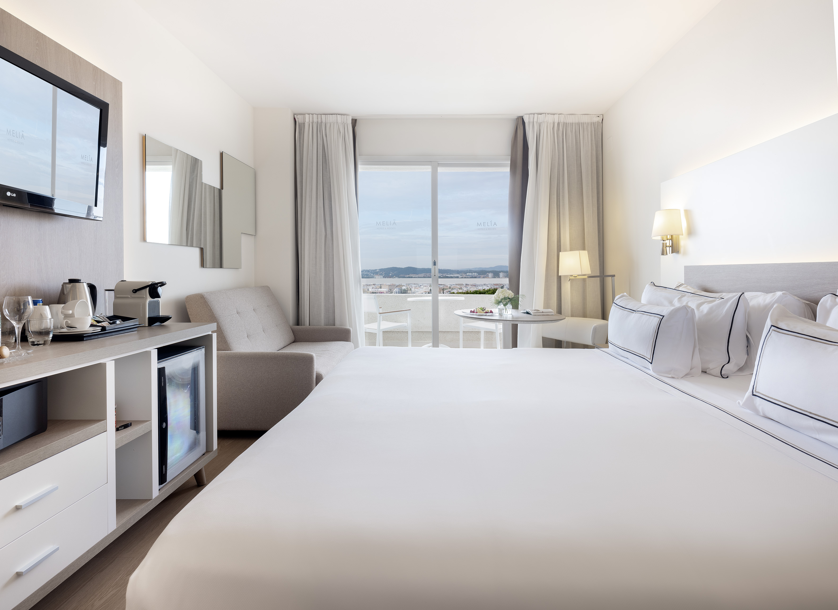 Modern resort room with white bed, balcony coastal view, TV, and minibar.