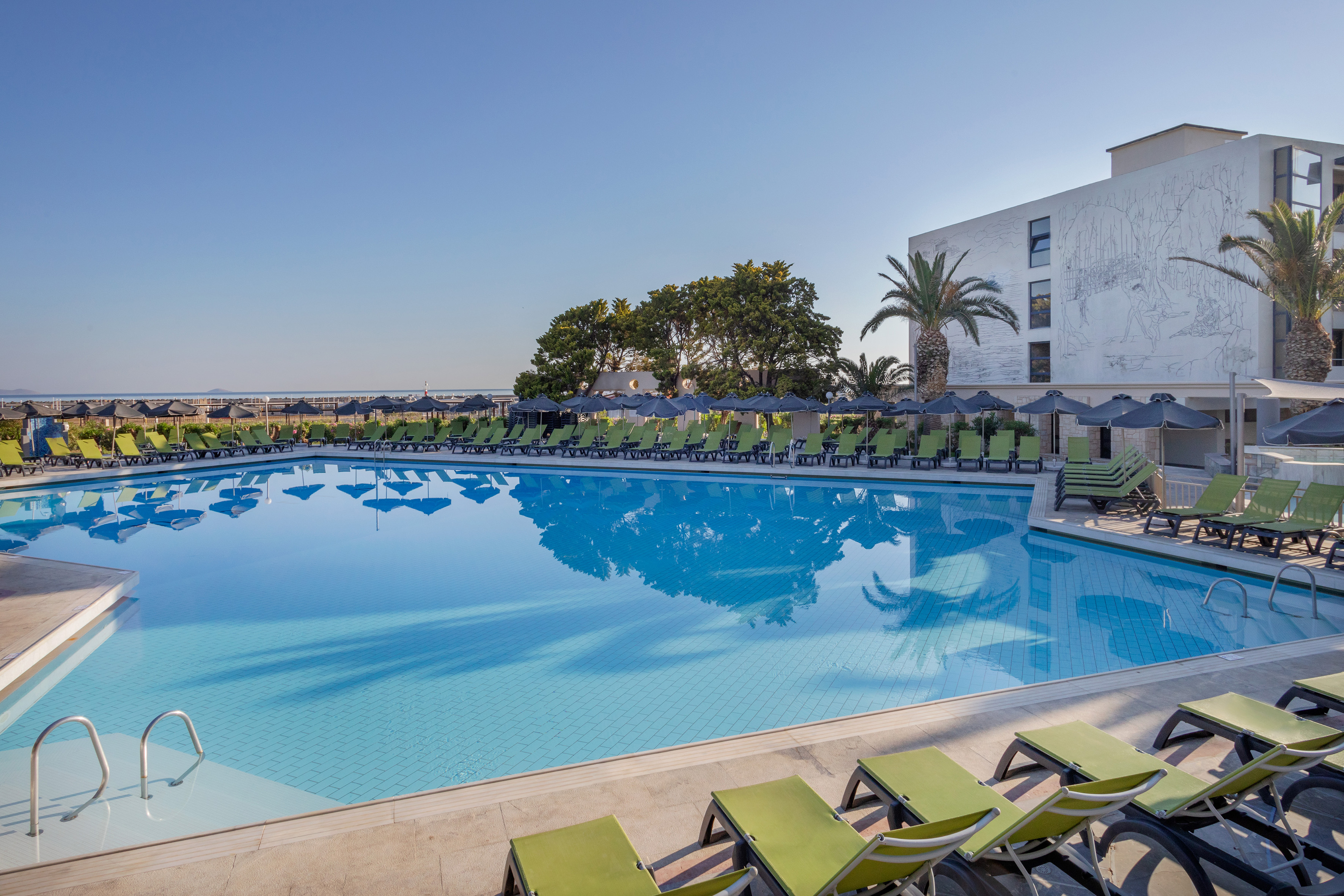 Outdoor hotel swimming pool with green loungers, umbrellas, and ocean view.