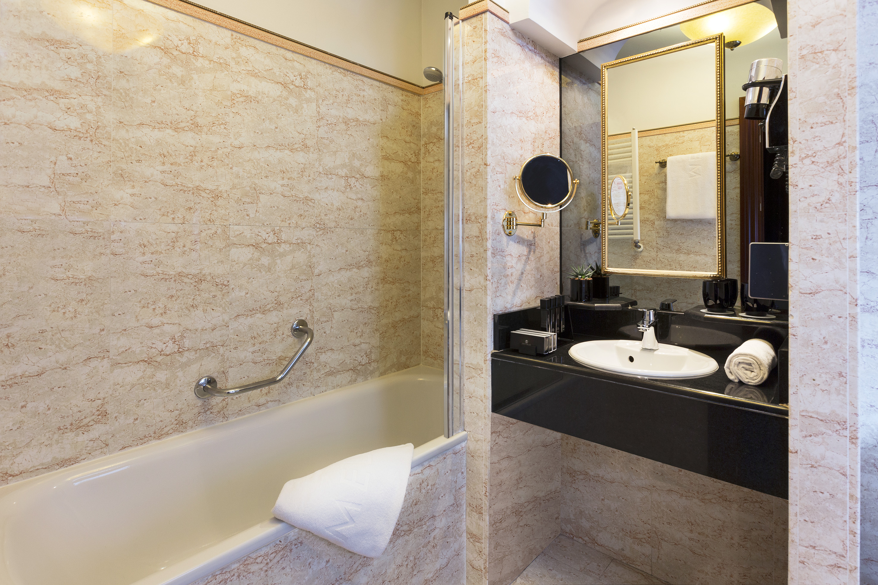 Hotel bathroom with marble walls, bathtub, black vanity, and mirror.
