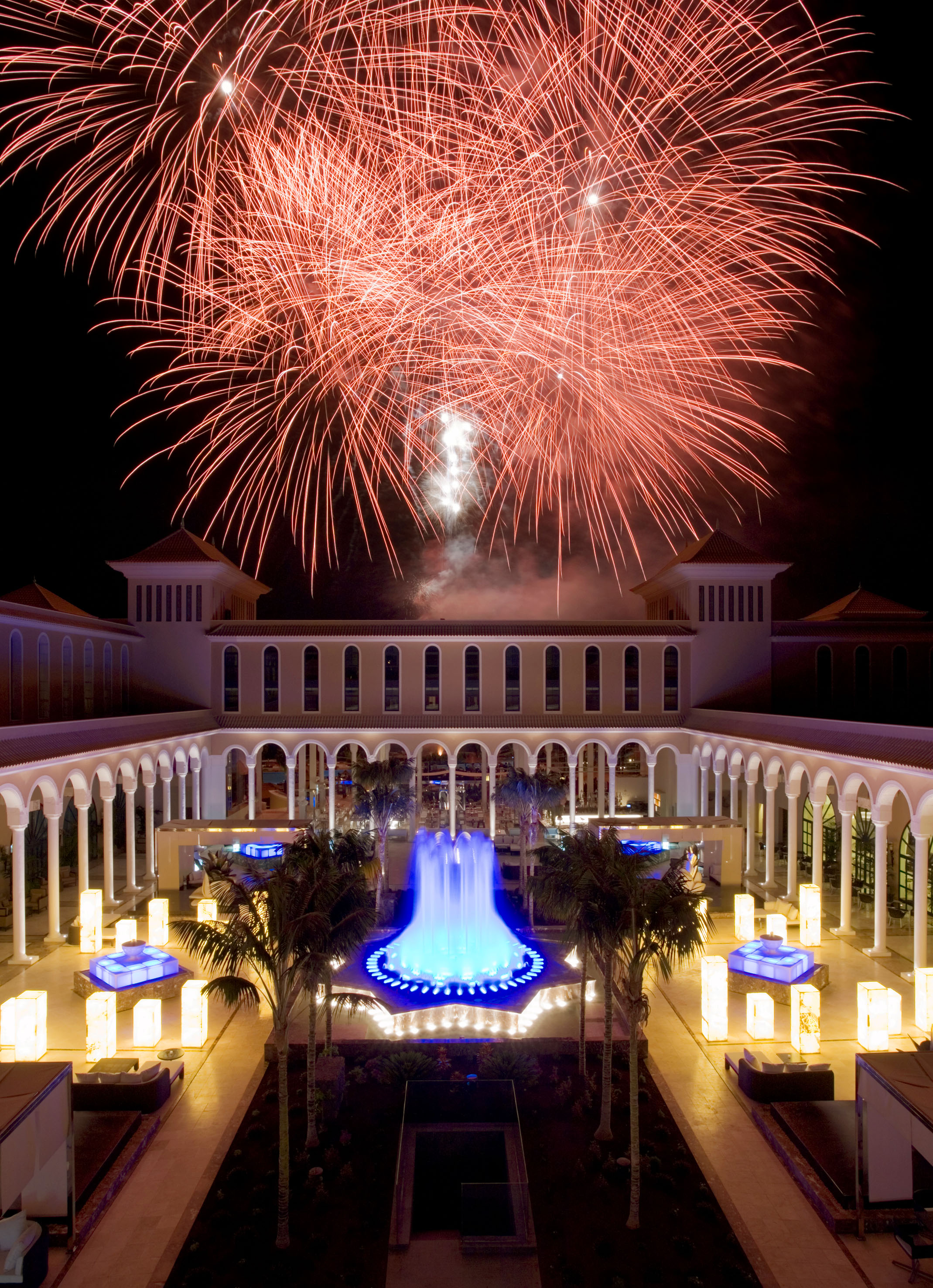 Red fireworks over a resort courtyard with a blue fountain and palm trees at night.