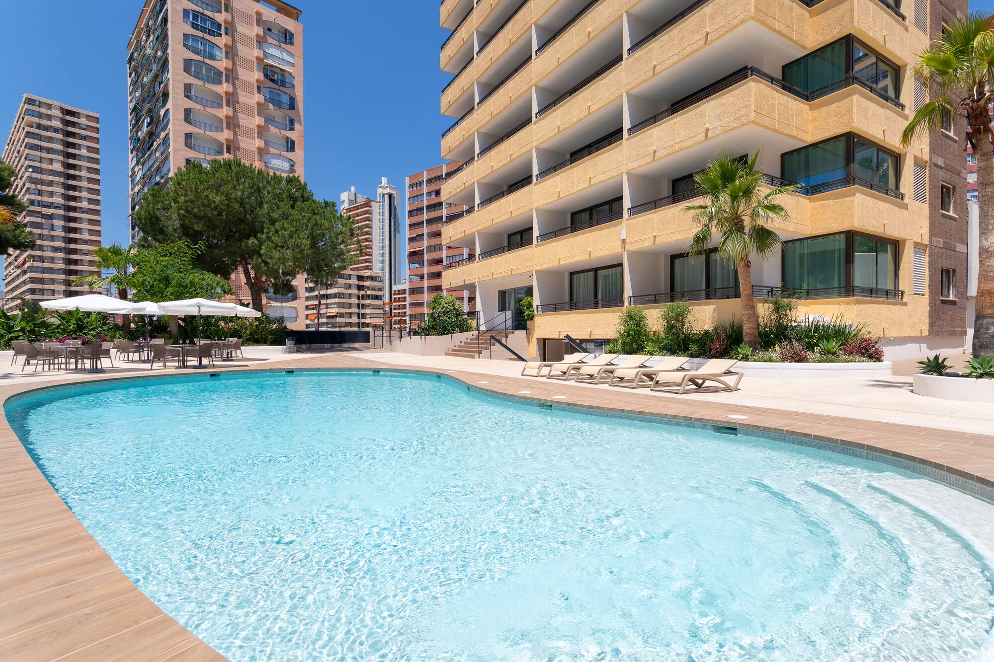 Outdoor hotel swimming pool with sun loungers, dining area, palm trees, and tall buildings.