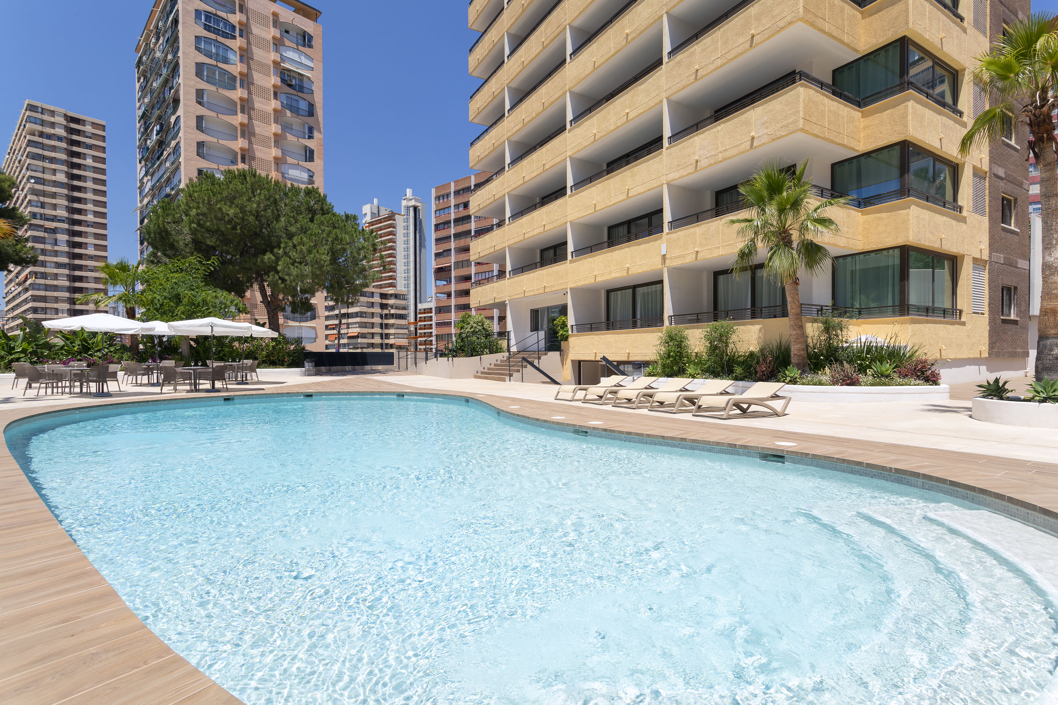 Outdoor hotel swimming pool with sun loungers, dining area, palm trees, and tall buildings.