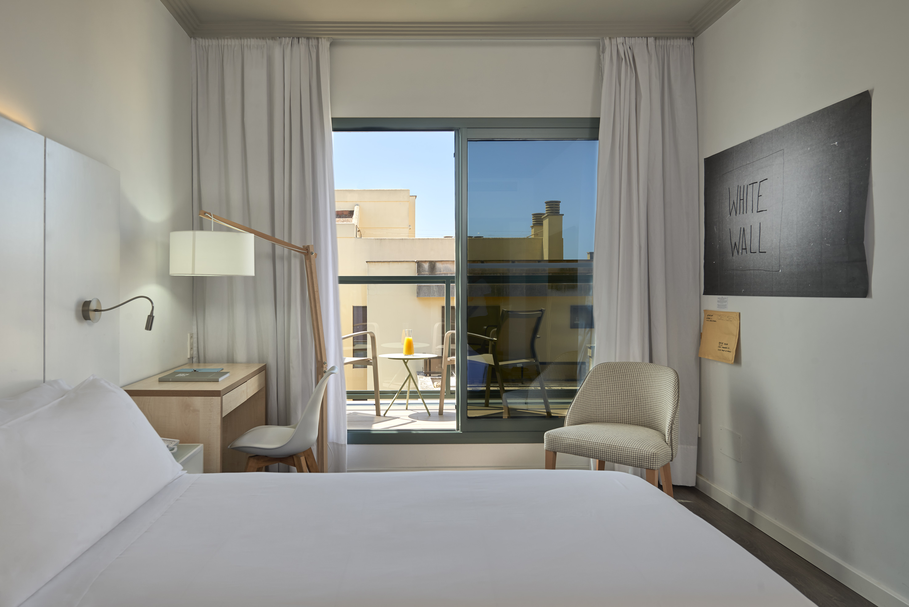 Modern hotel bedroom with white bed, desk, and balcony city view.