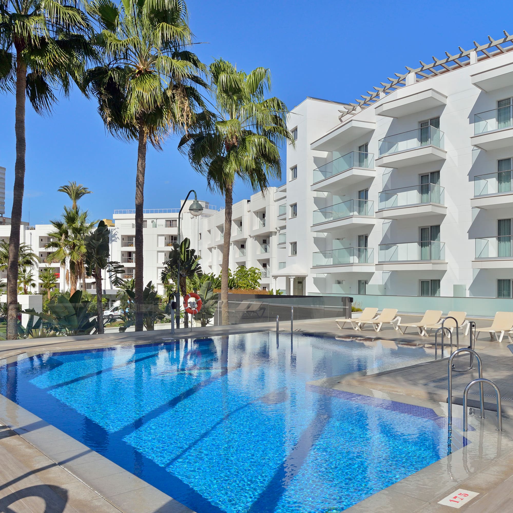 Sunny resort swimming pool with modern white hotel, balconies, and palm trees.