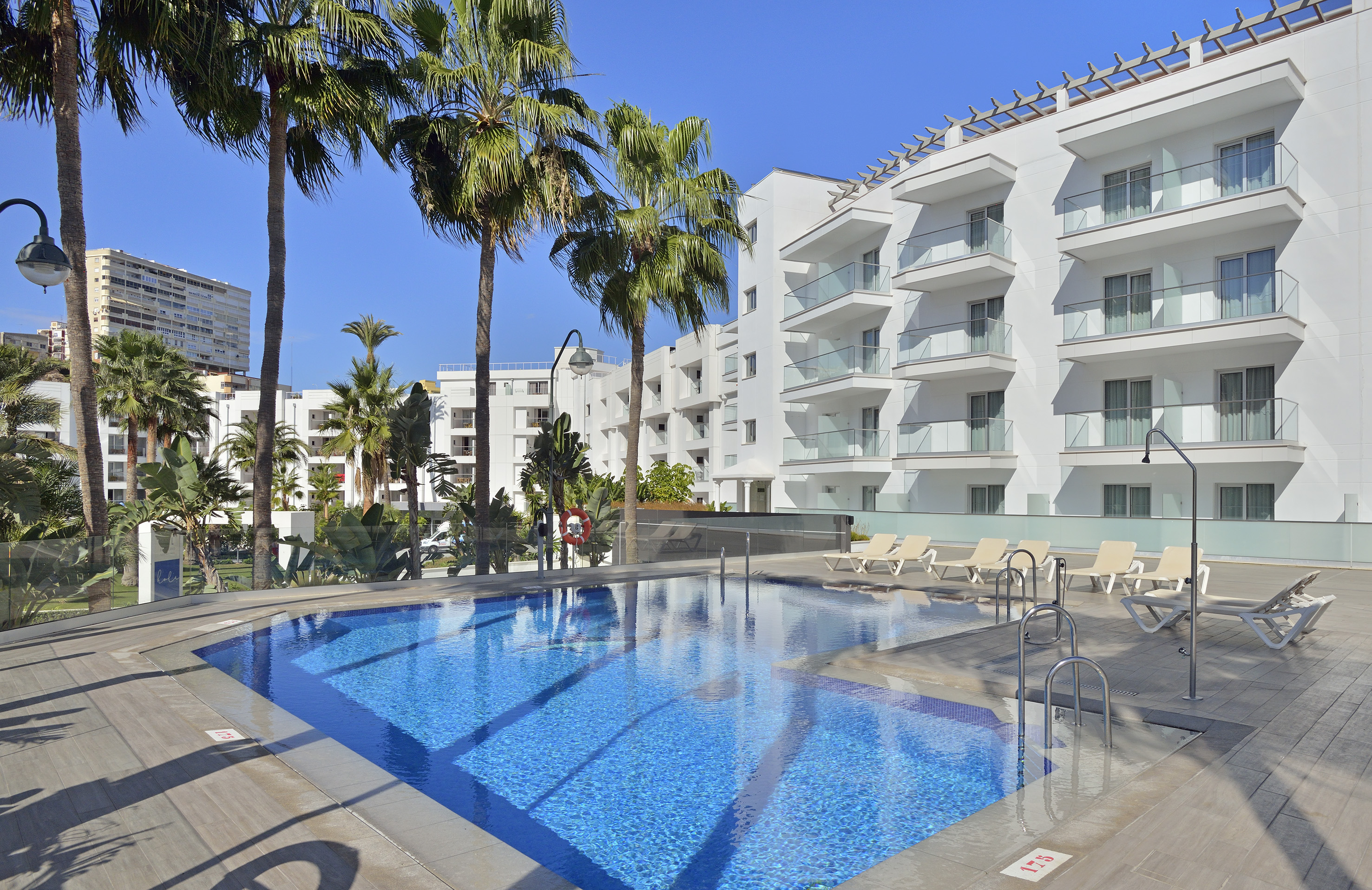 Modern resort swimming pool with white buildings, balconies, and palm trees.
