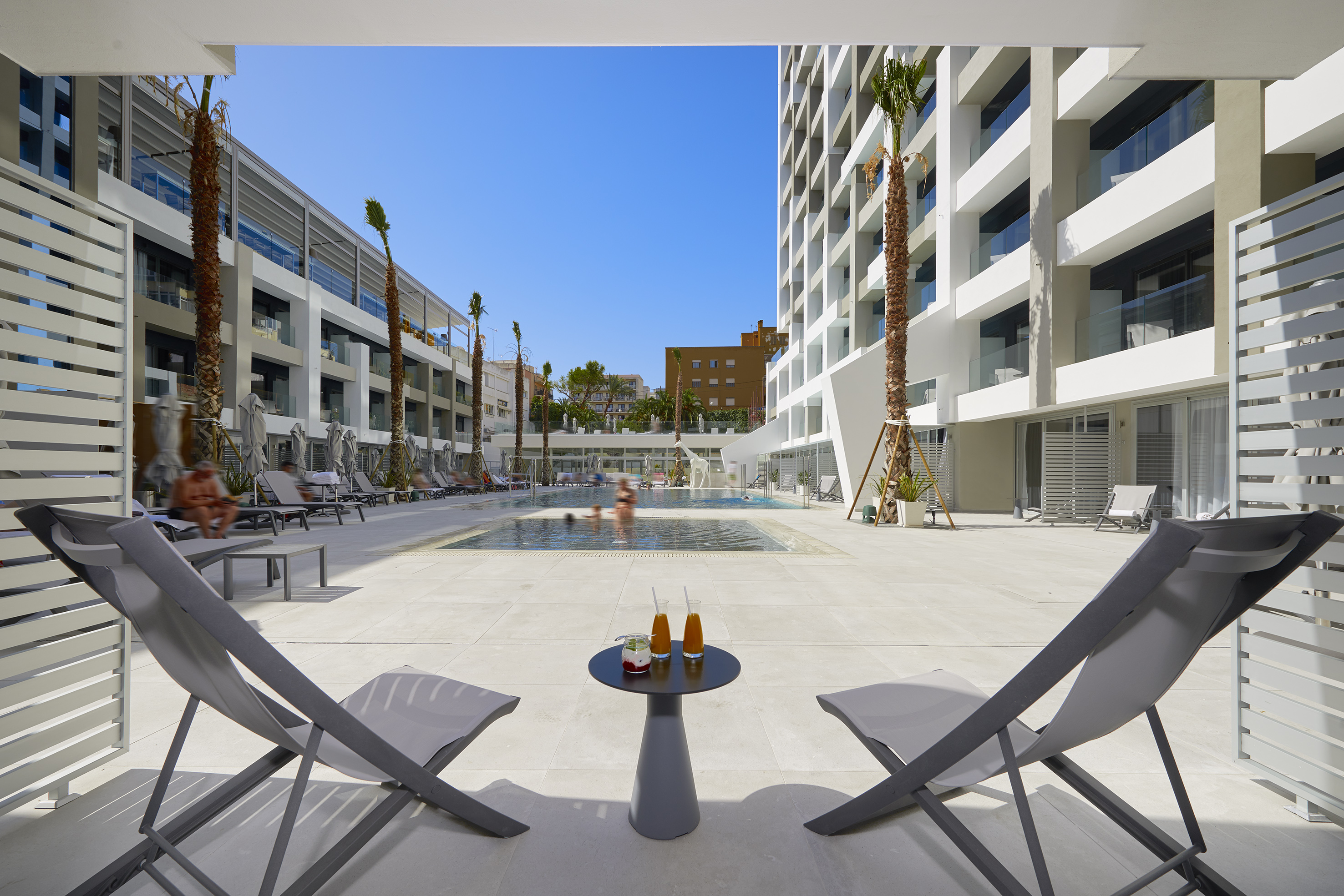 Resort pool deck with lounge chairs, cocktails, palm trees, and modern hotel.