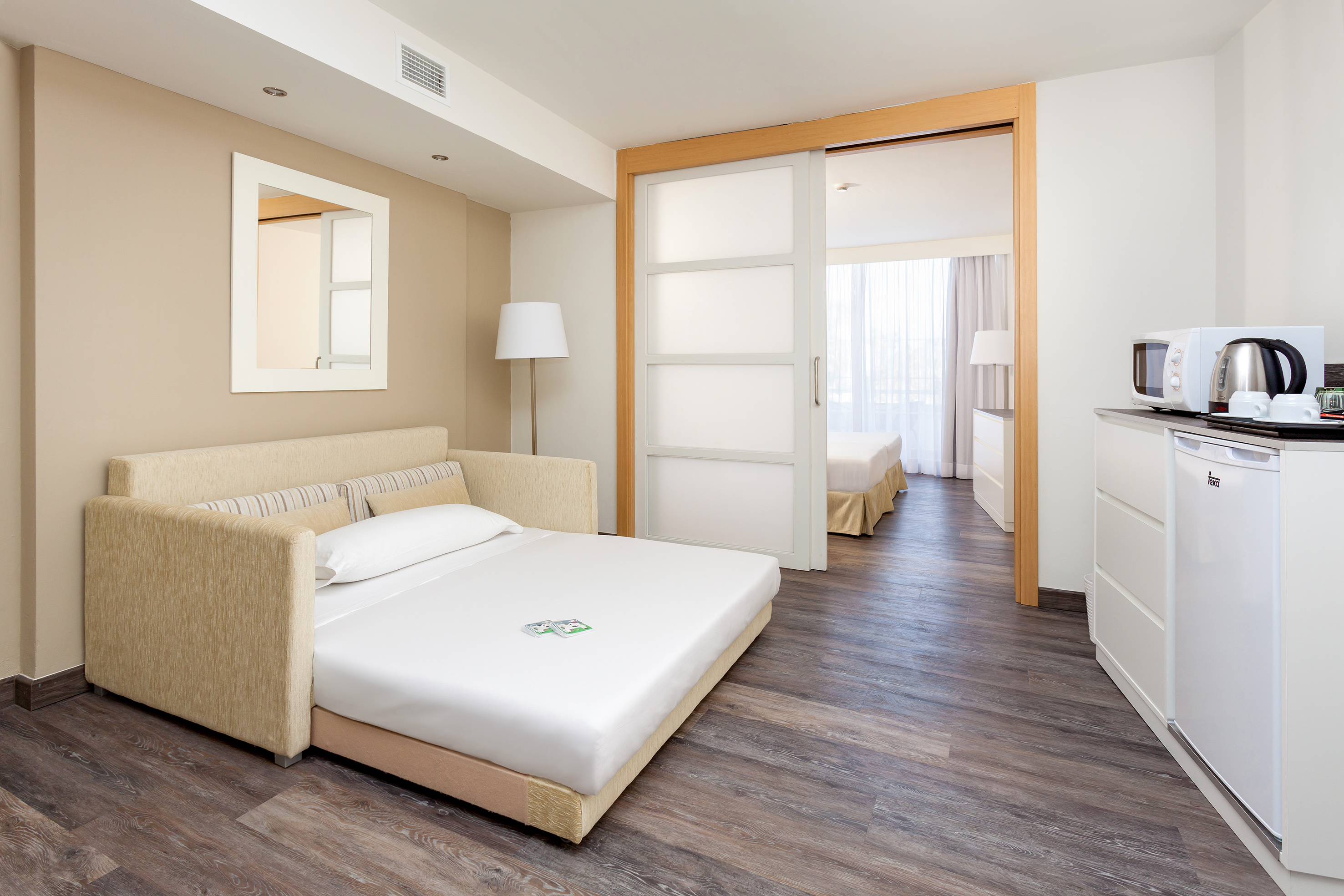 Modern hotel suite with extended sofa bed, separate bedroom, and kitchenette.