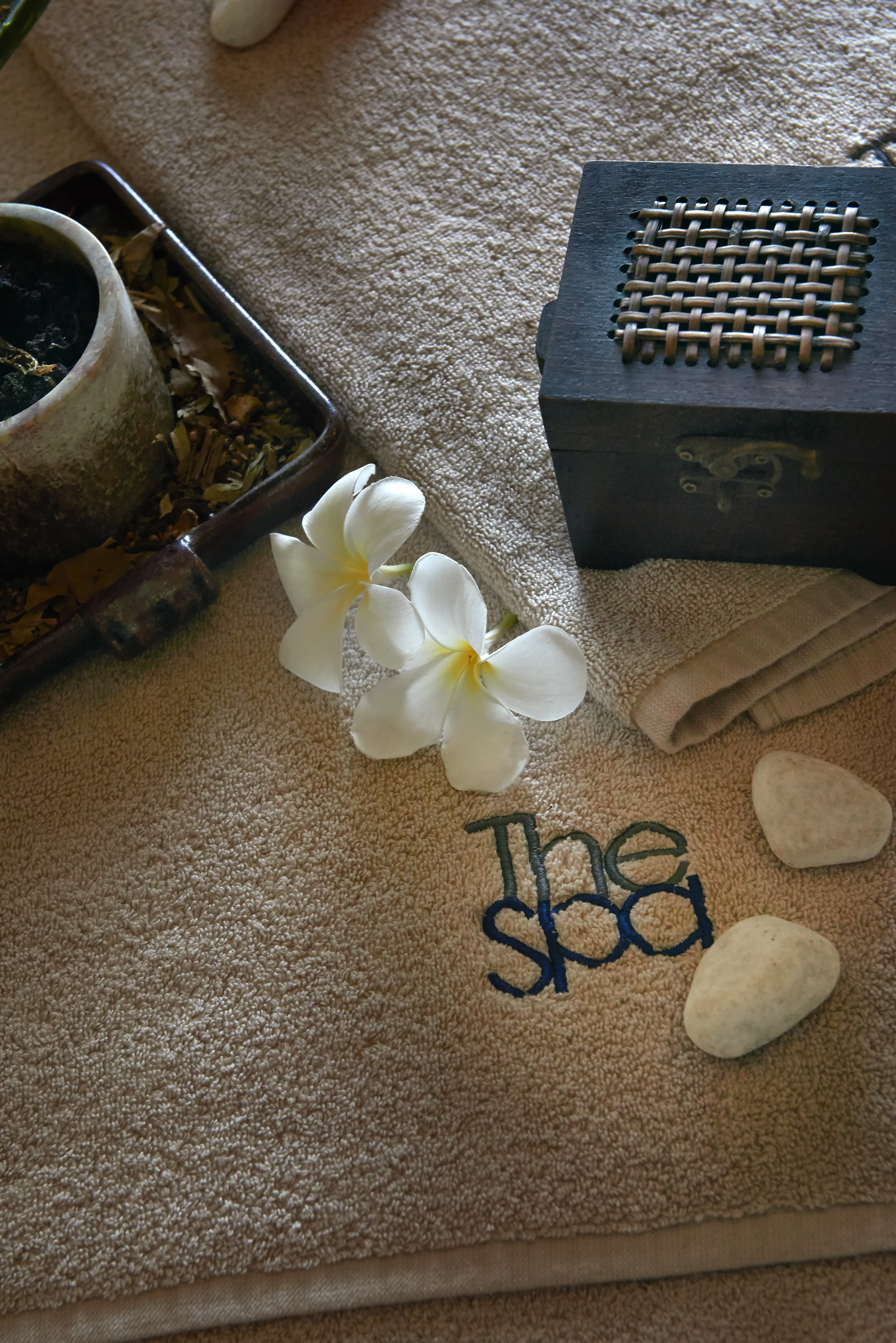 Beige spa towel with "The Spa" embroidery, white plumeria flowers, and smooth stones.