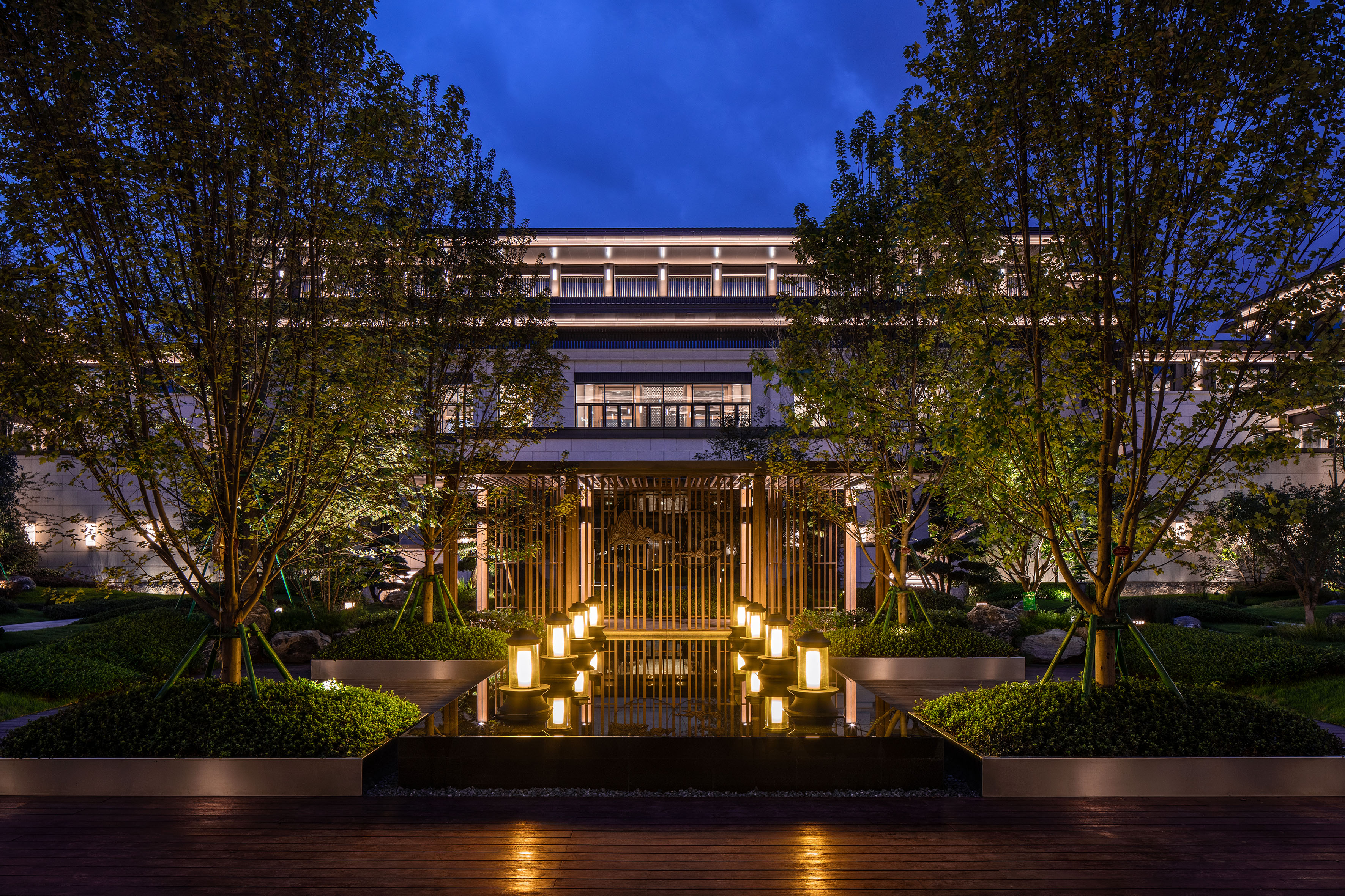Illuminated modern building, trees, and reflective water feature with lanterns at dusk.