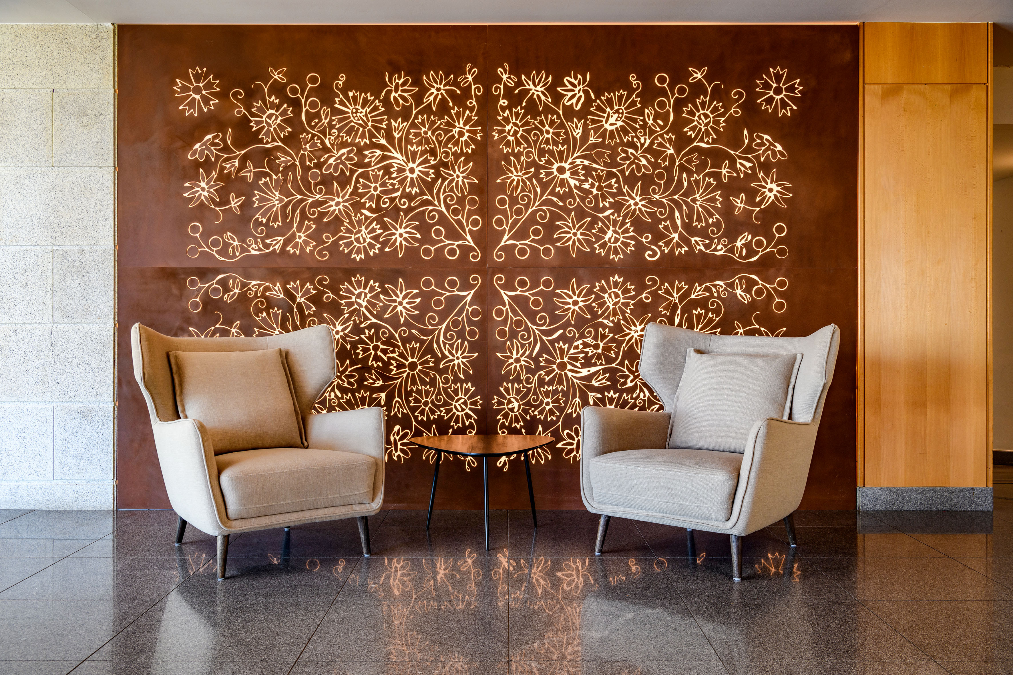 Modern lounge with beige armchairs, wooden table, and backlit floral wall art.