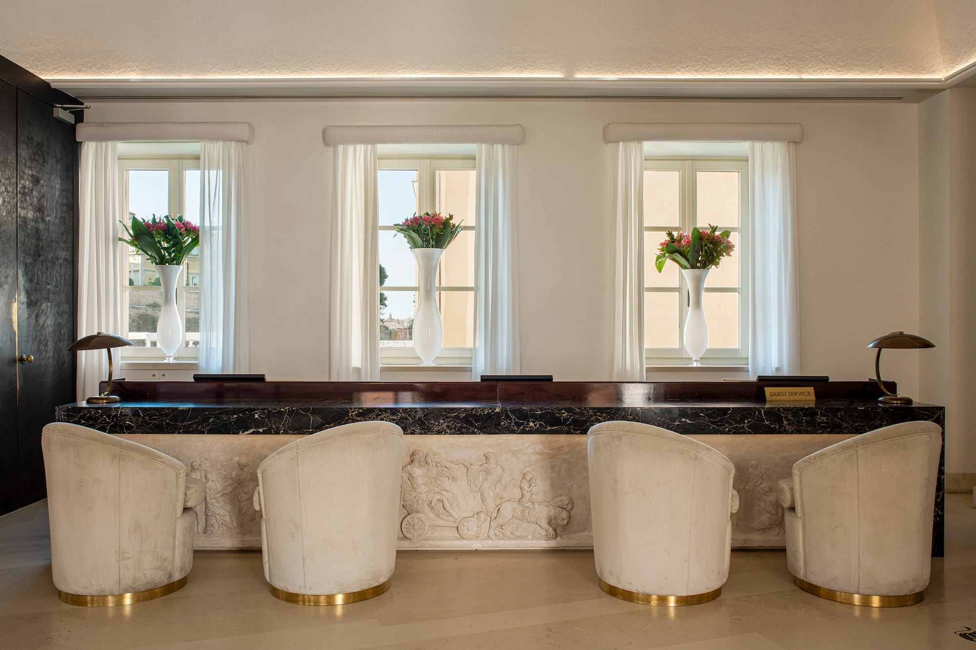 Luxury hotel reception desk with carved marble, cream chairs, and flowers.