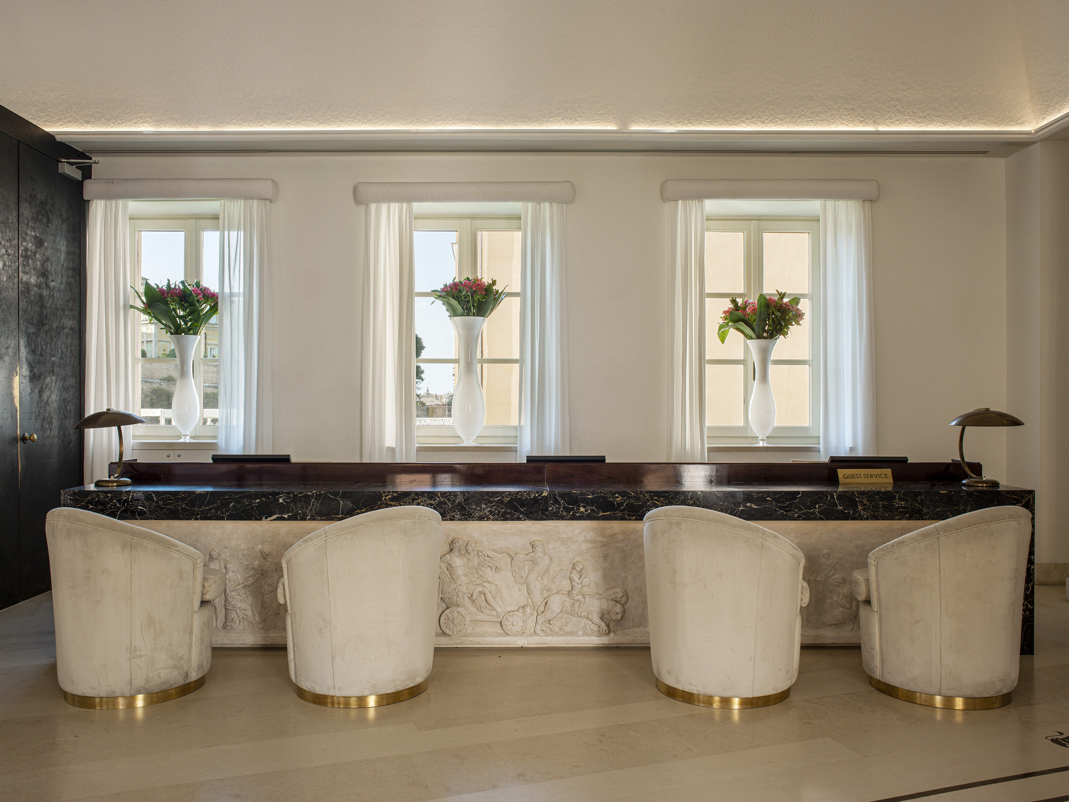 Luxury hotel reception desk with carved marble, cream chairs, and flowers.