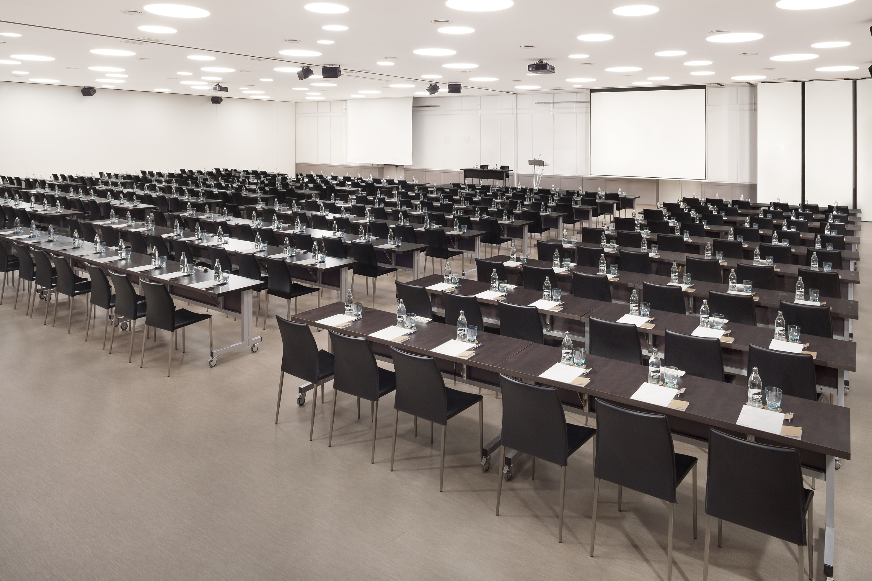 Empty modern conference room with tables, chairs, water, and projection screens.
