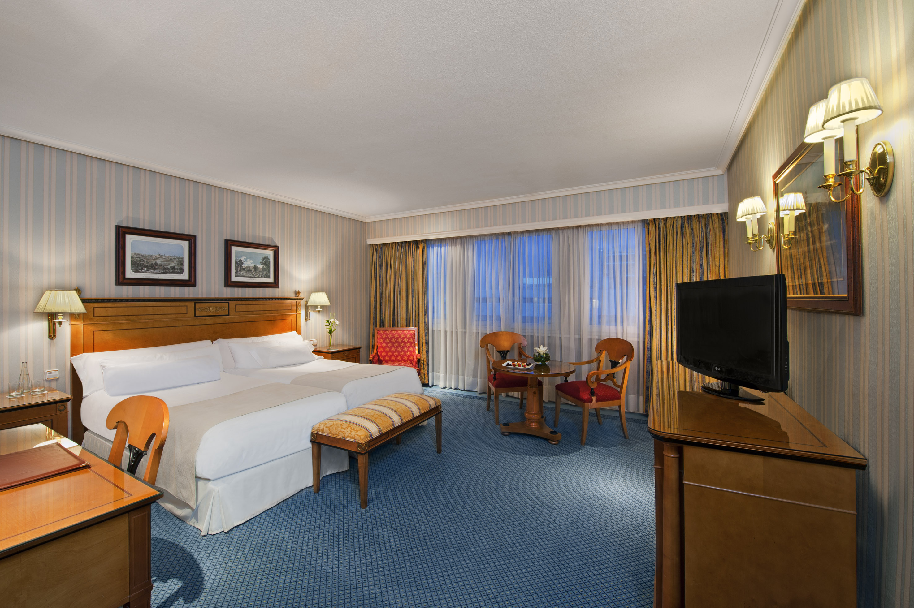 Classic hotel bedroom with two beds, blue carpet, wooden furniture, and striped walls.