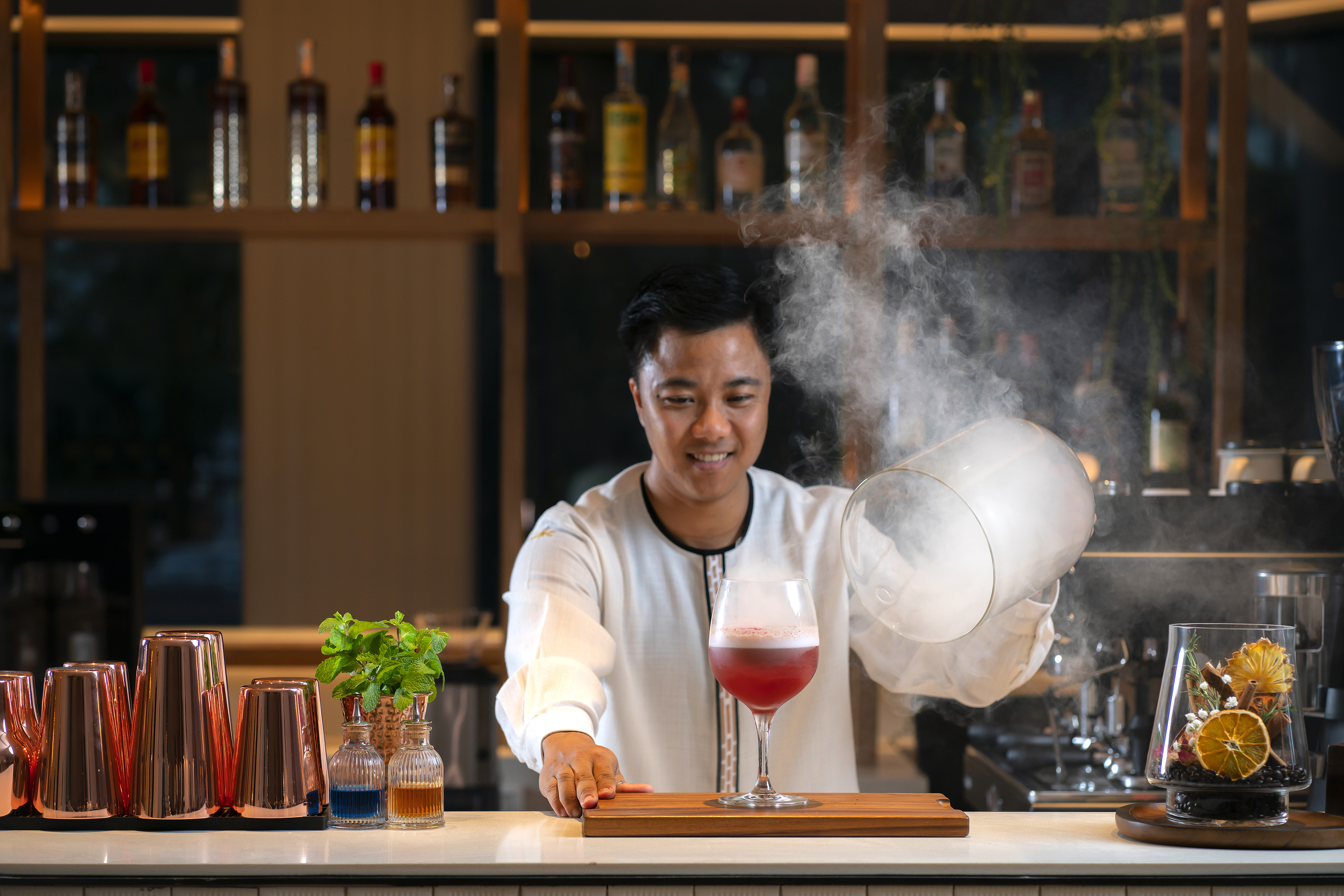 Mixologist smiling, smoking a red cocktail under glass dome at a bar.