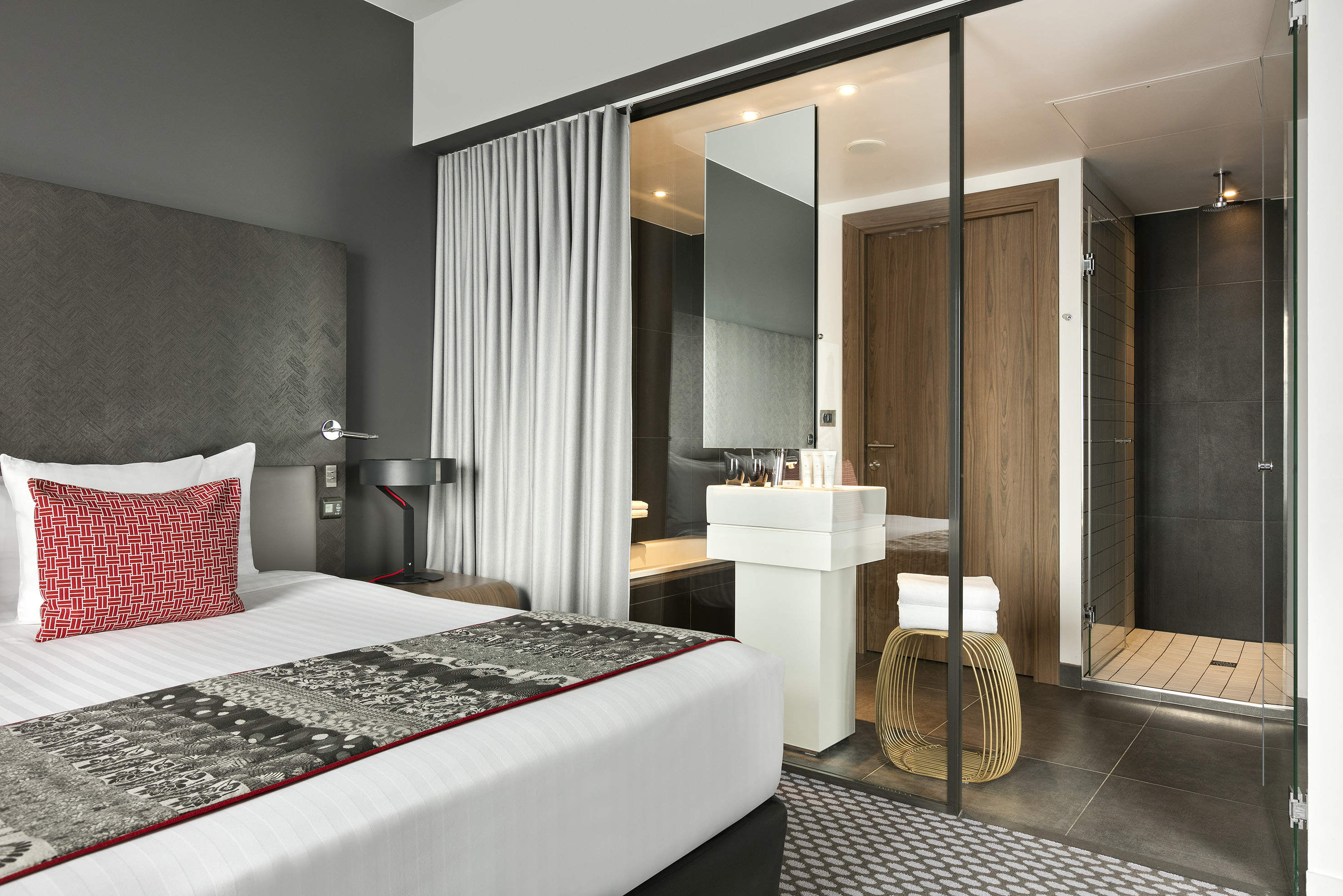 Modern hotel bedroom with red pillow, glass bathroom, vanity, and shower.