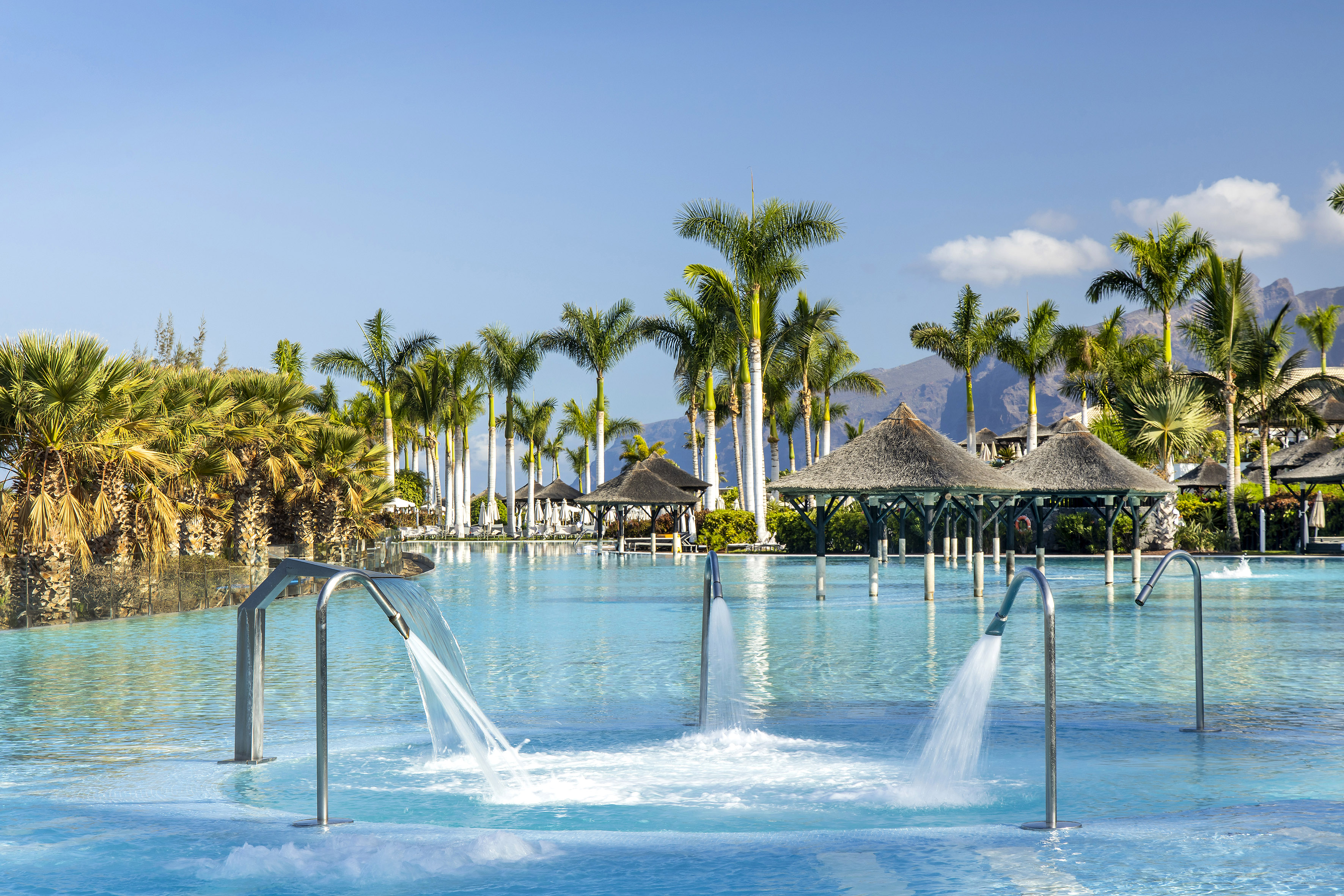 Resort swimming pool with water jets, palm trees, straw huts, and mountains.
