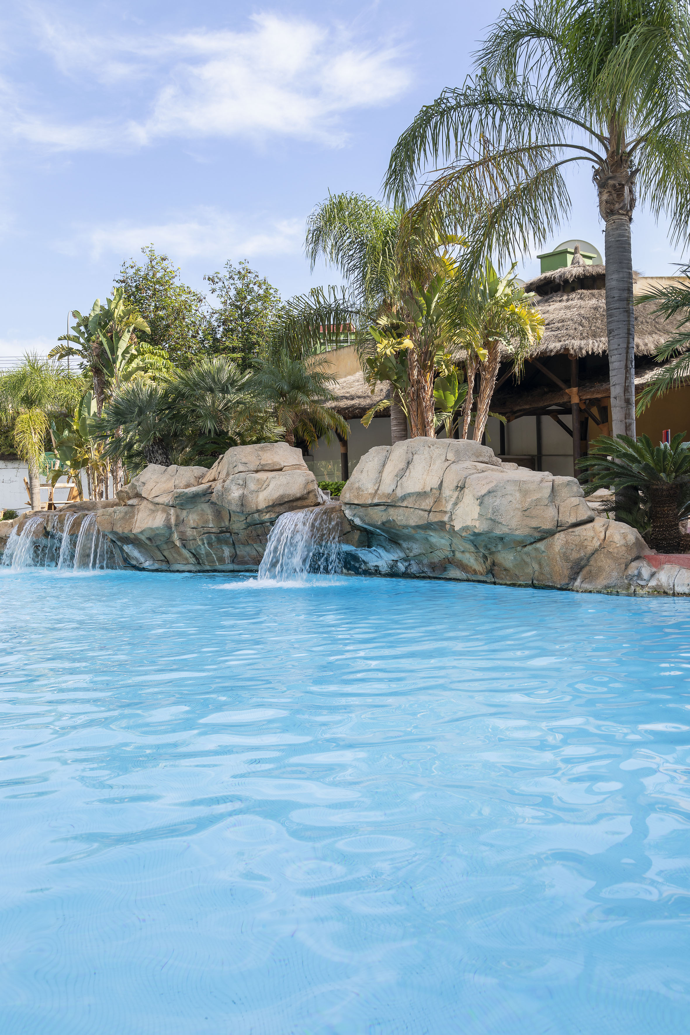 Outdoor pool with waterfalls, tropical palm trees, and thatched roof cabana.