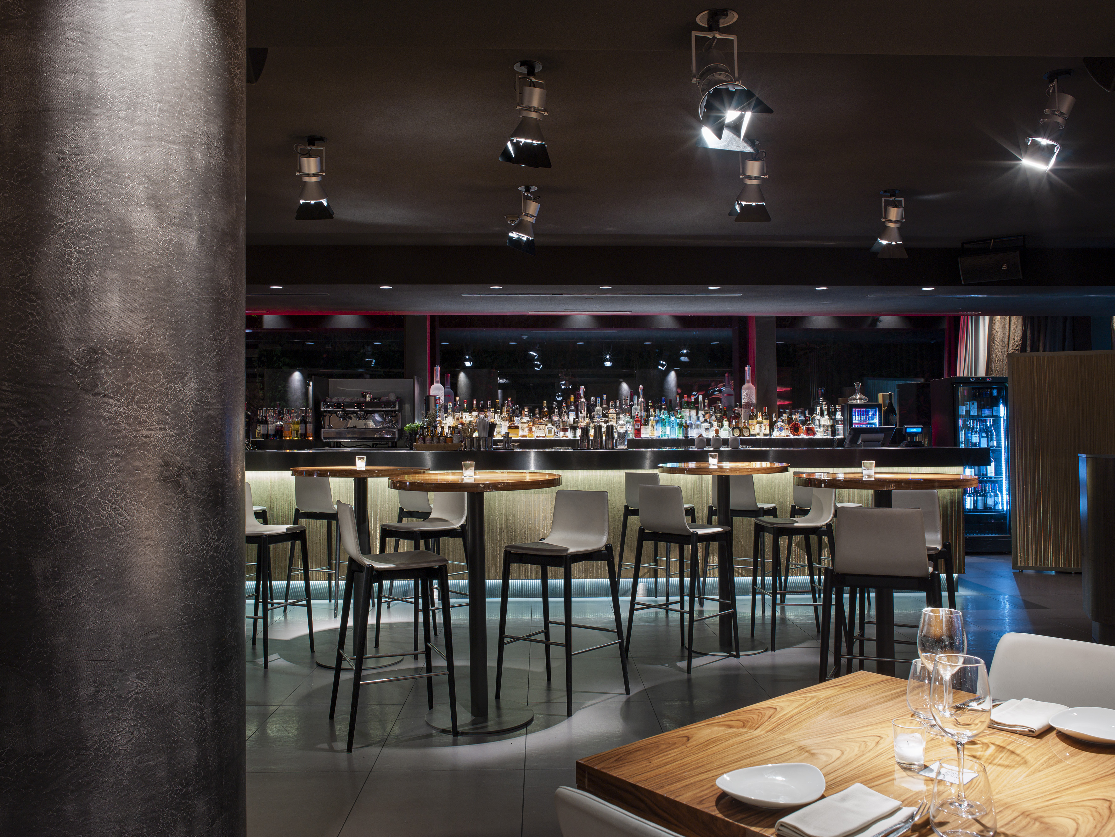 Modern dark bar interior with backlit liquor bar and high-top tables.