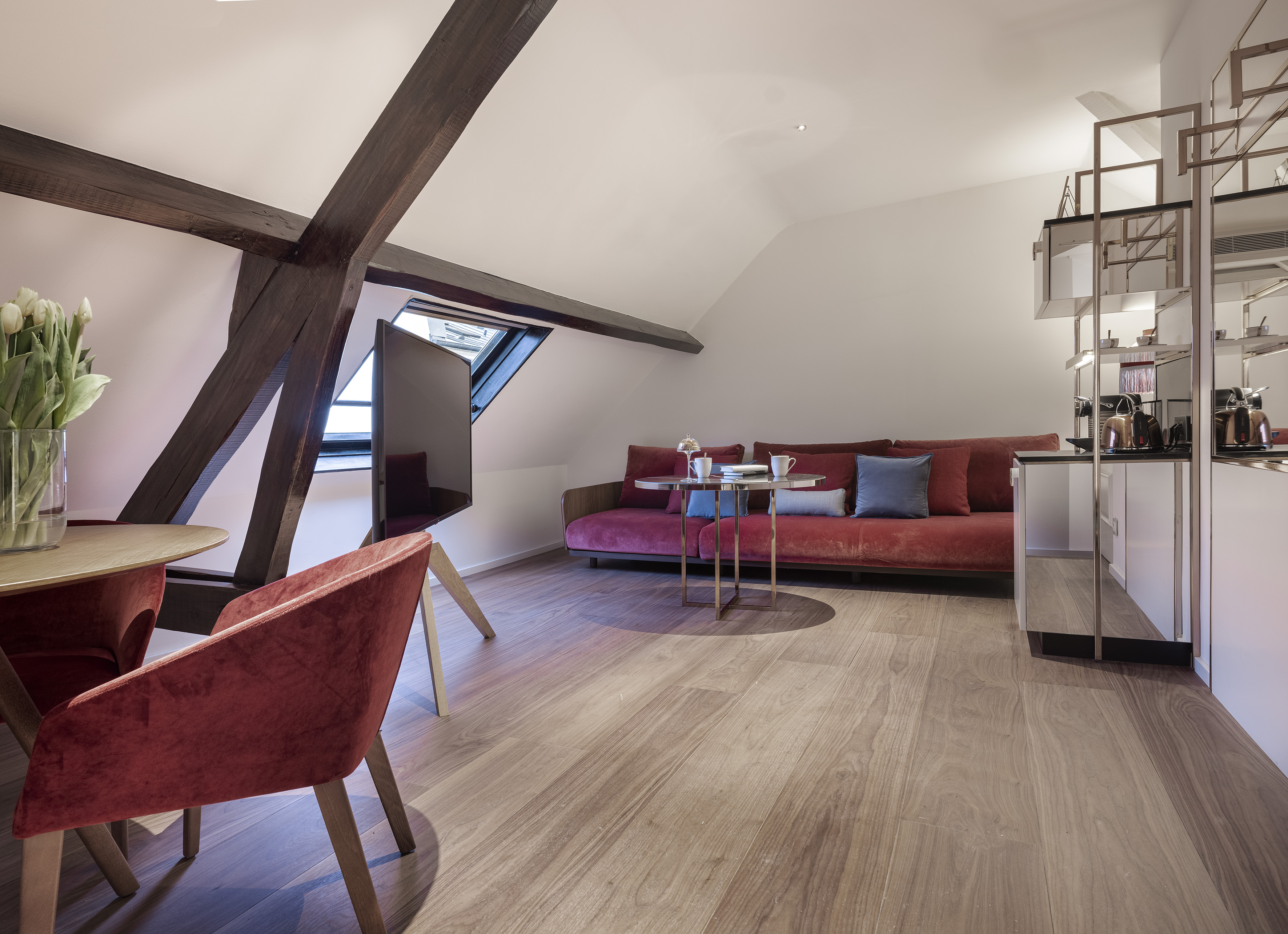 Attic apartment living space with red sofa, wooden beams, skylight, and kitchenette.