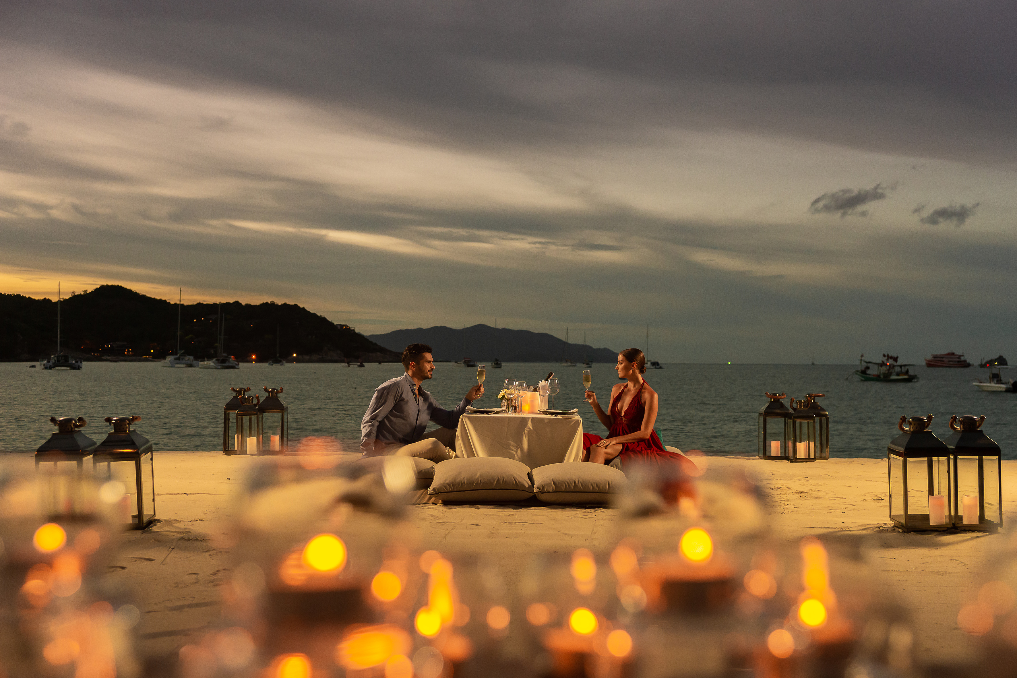 a man and woman sitting at a table with wine glasses on the beach