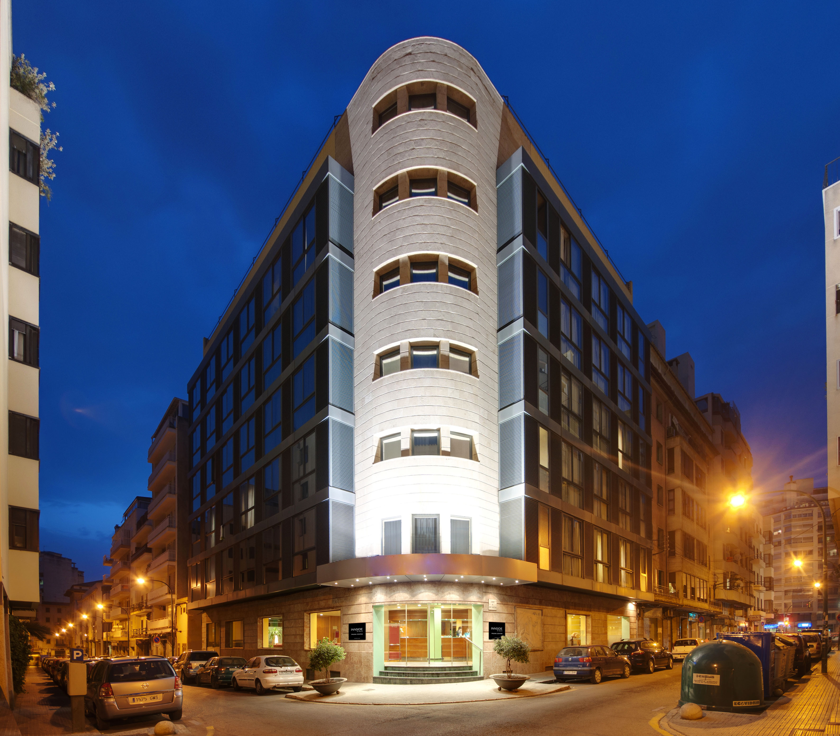 Modern curved building facade at dusk, city street with parked cars and lights.